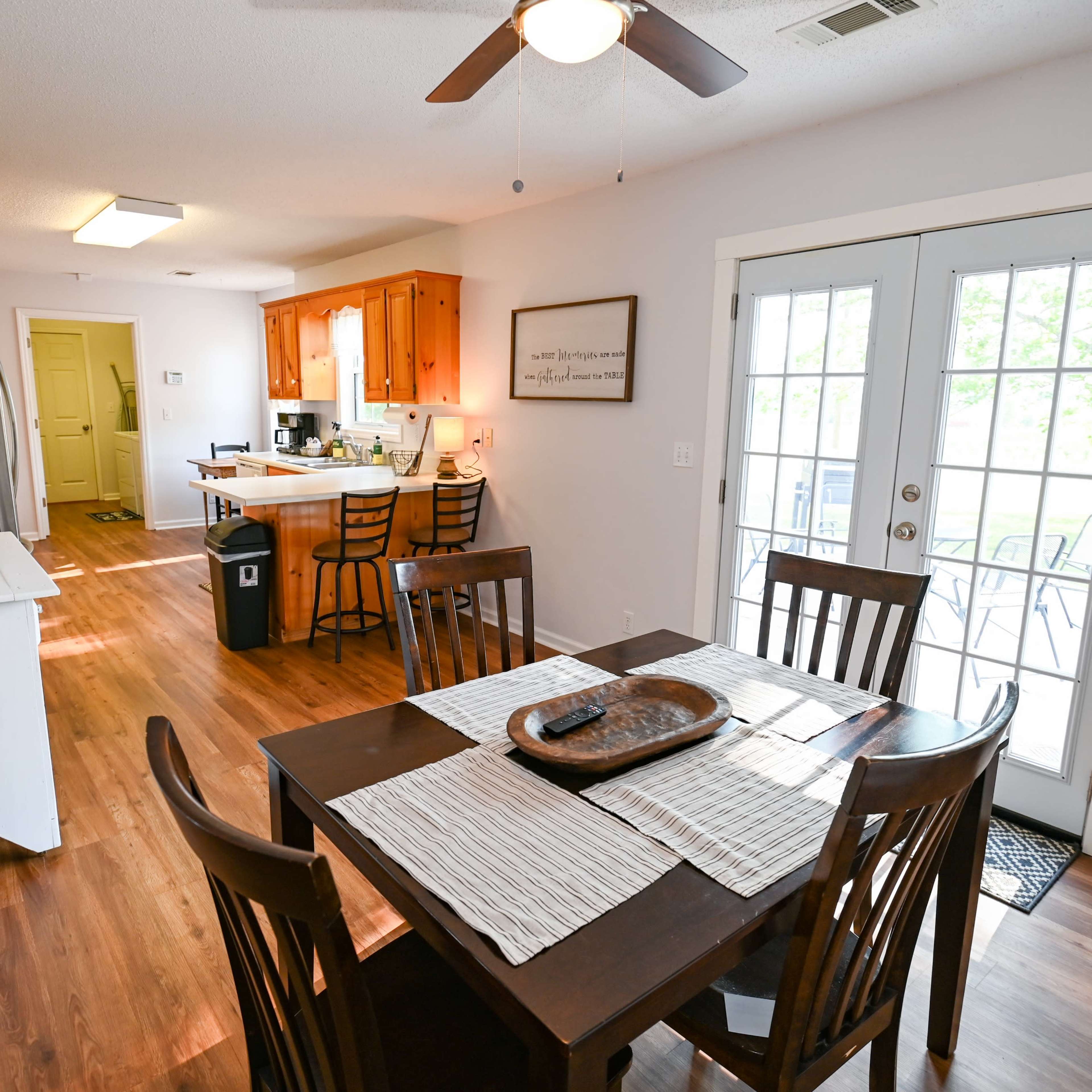 A dining area with a wooden table and chairs is adjacent to a kitchen, illuminated by natural light coming through double doors leading to an outdoor space.