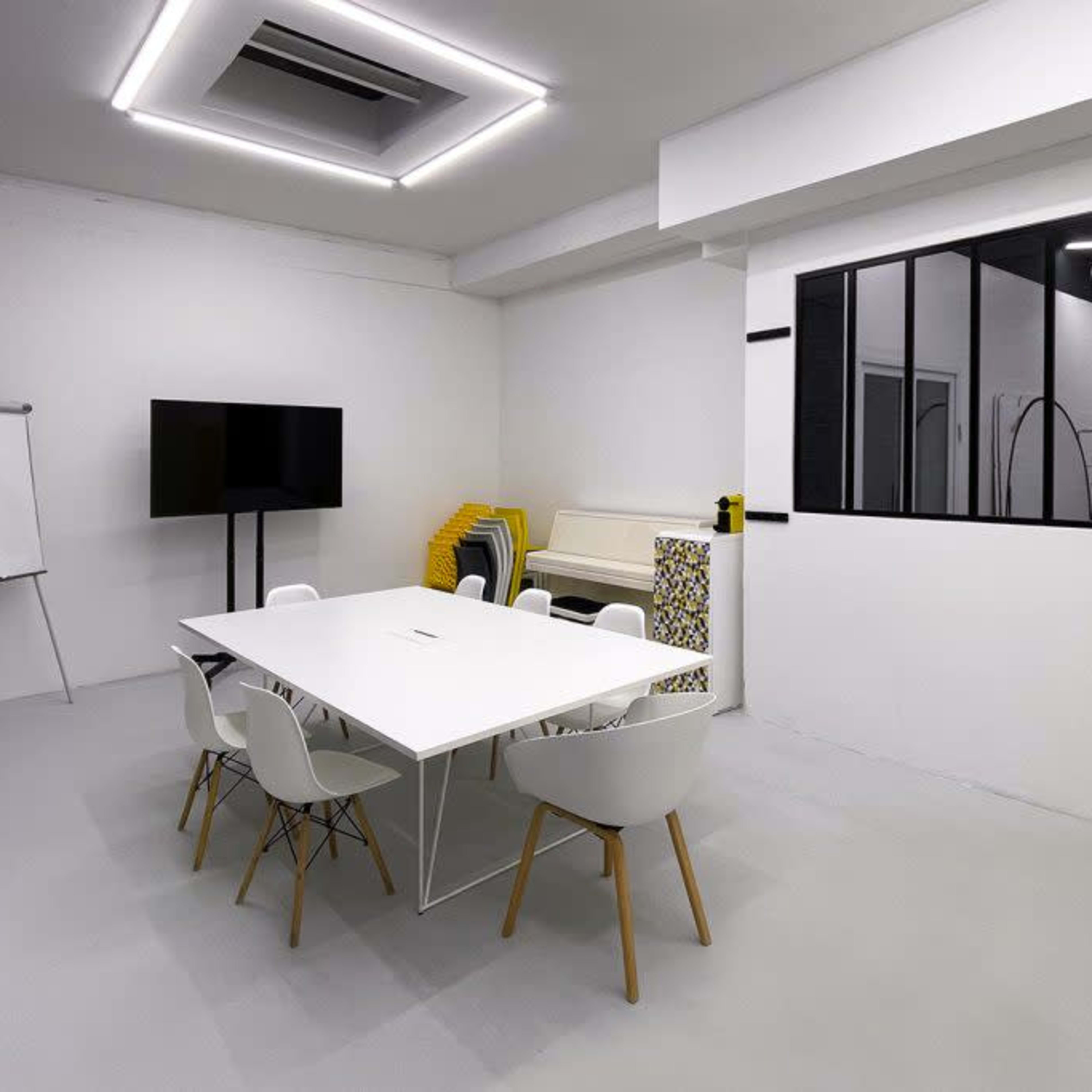 A modern meeting room features a large white table surrounded by white chairs, a television screen, a yellow door, and a wall window.