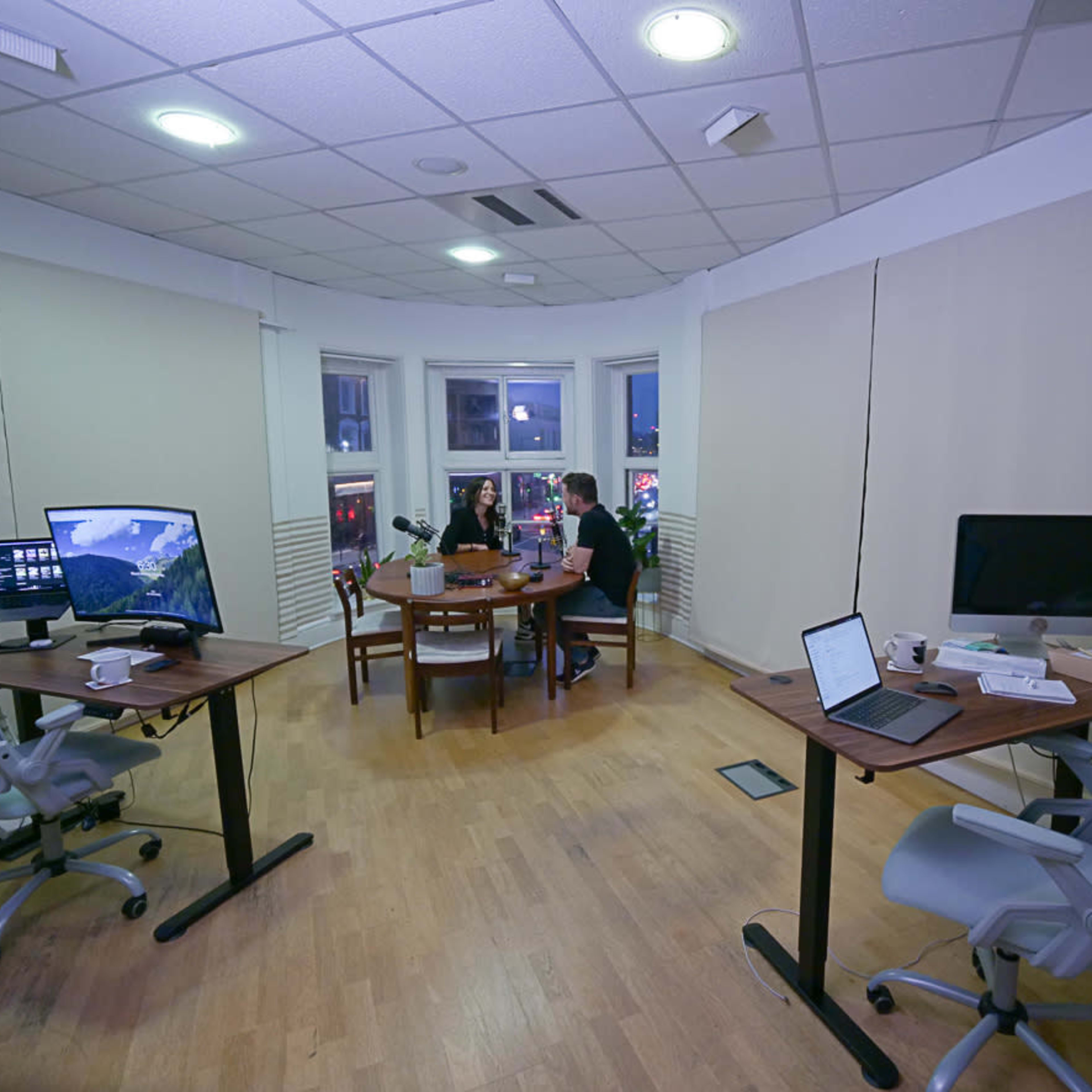 A modern office space with two workstations, a small dining table, and large windows overlooking a city street at night.
