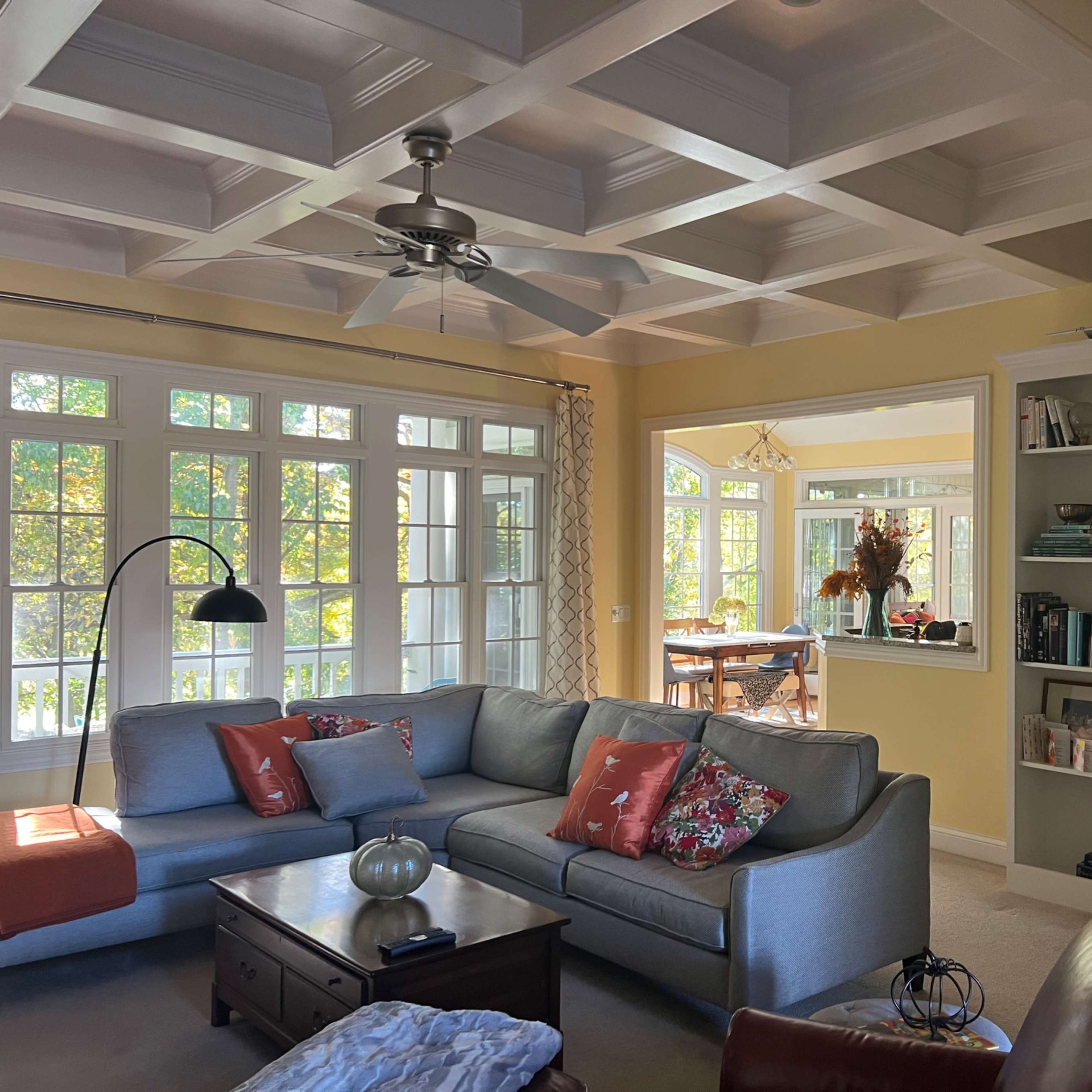 A bright living room with a coffered ceiling, a large gray sofa, a wooden coffee table, and a built-in bookshelf filled with books.