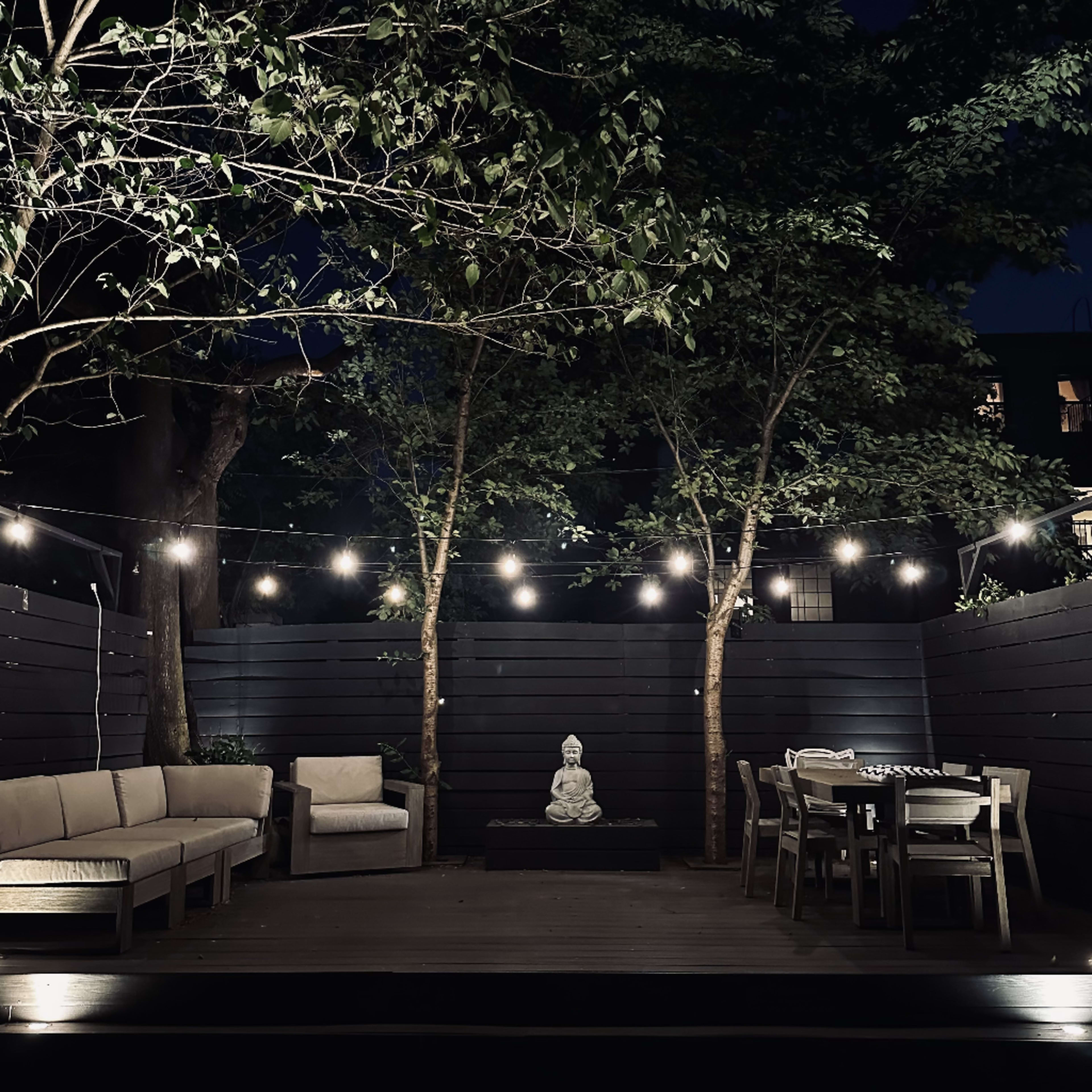 A well-lit outdoor seating area features a stone statue surrounded by trees, with string lights overhead illuminating a sectional sofa and dining table.