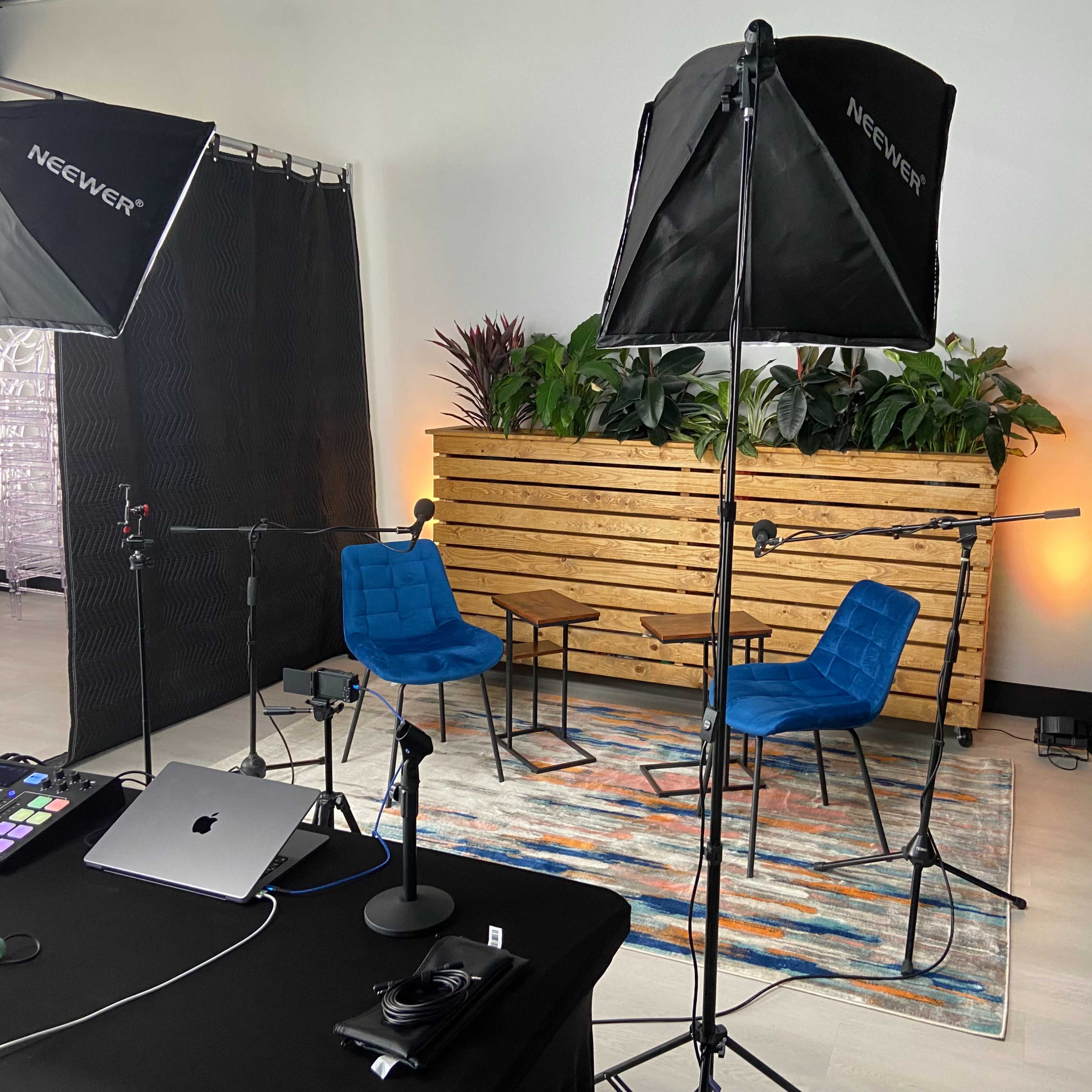 A prepared video recording setup featuring two blue chairs, a small wooden table, lighting equipment, and a laptop on a black table, with plants in the background.