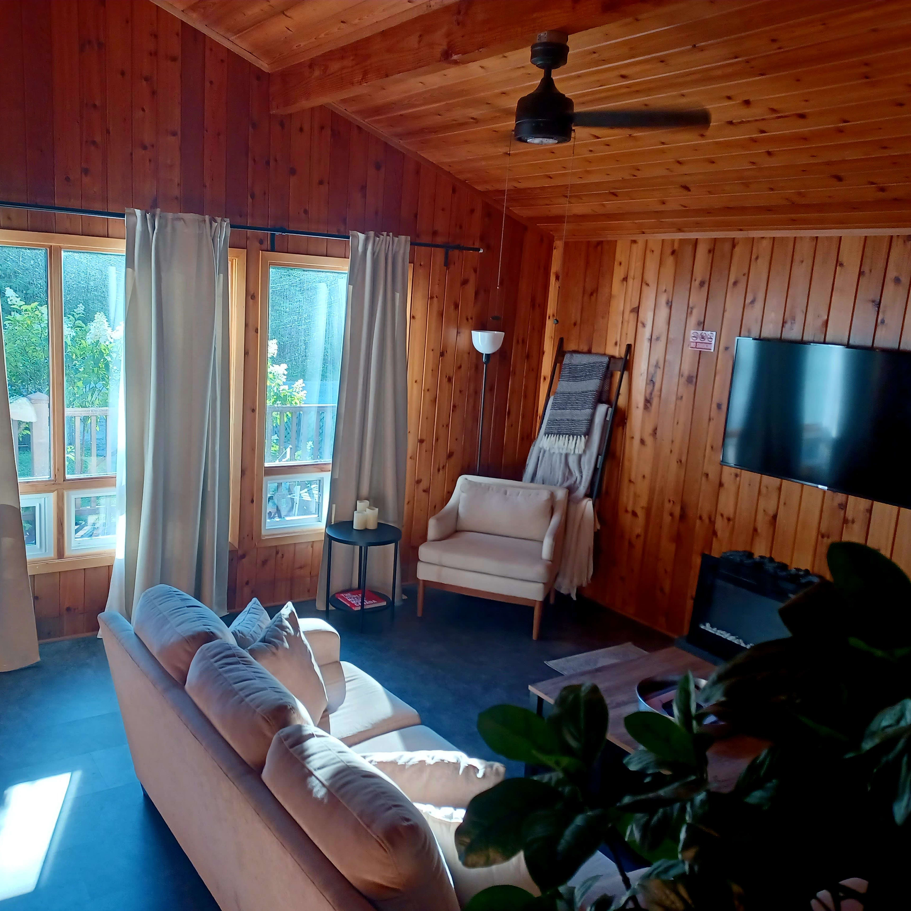 A cozy room with wooden walls, large windows, a light-colored sofa, and a television mounted on the wall.