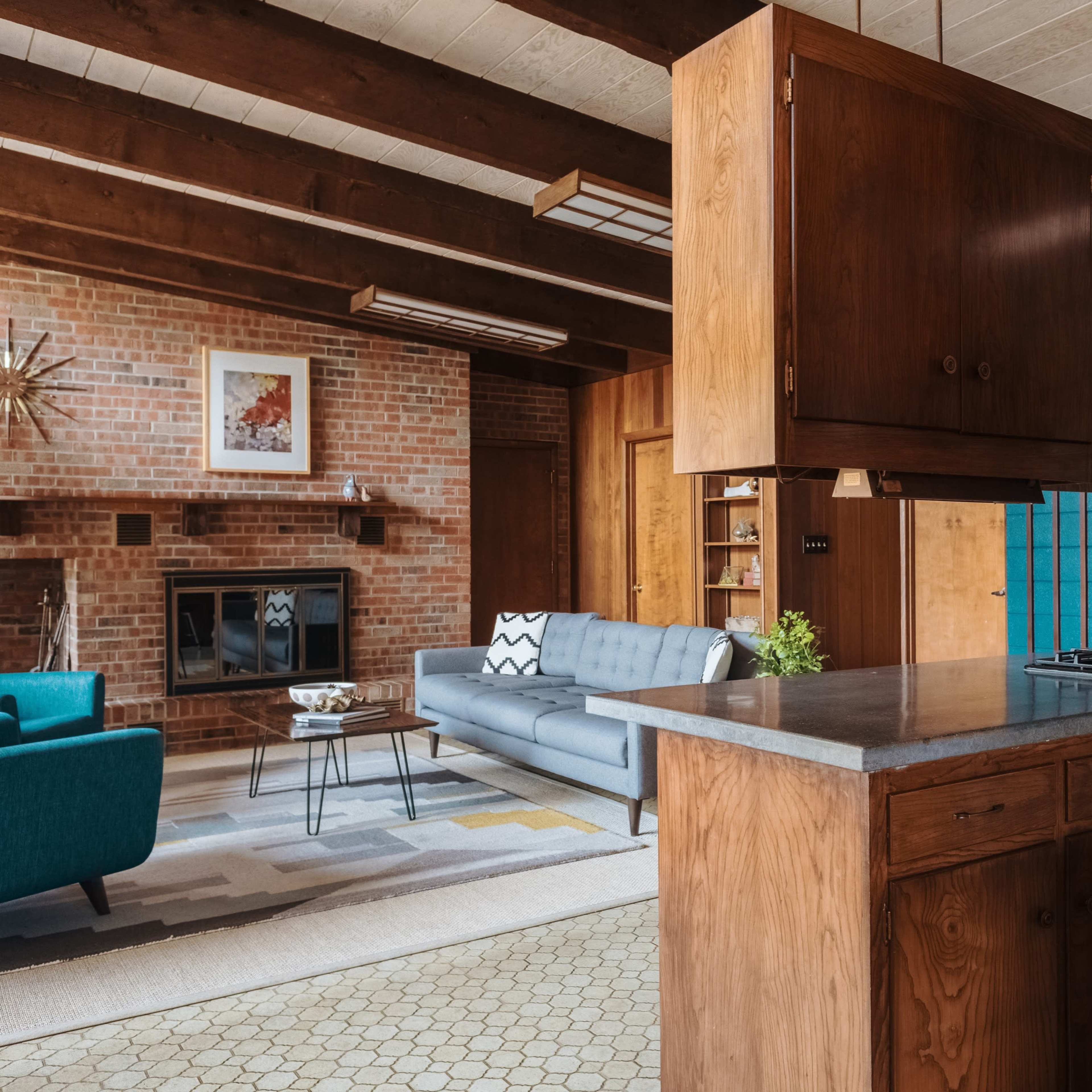 A modern open-concept kitchen and living area with wooden cabinetry, a granite countertop, and a cozy seating arrangement featuring a gray sofa and blue armchairs.