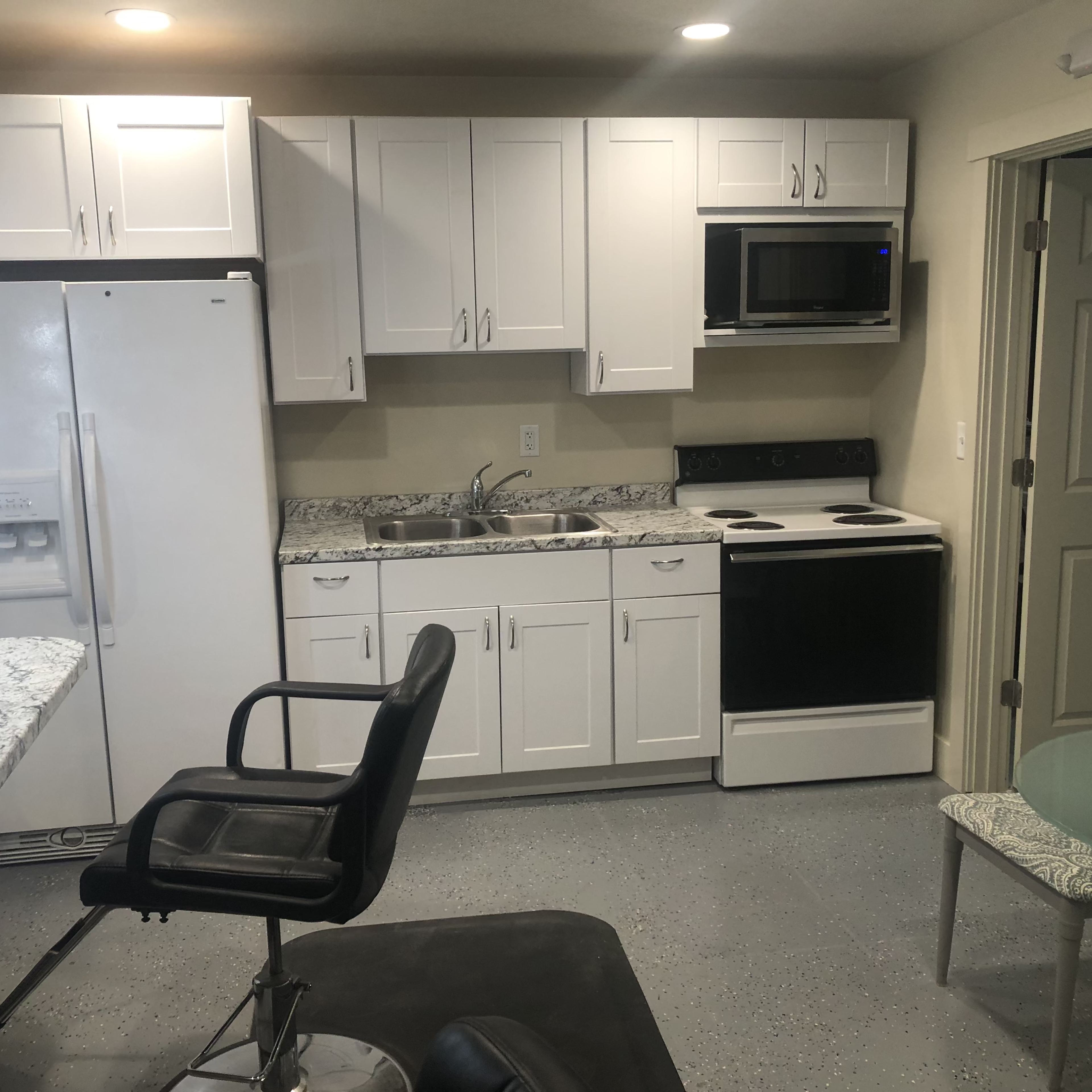 A compact kitchen with white cabinets, a stainless steel sink, a microwave, an electric stove, and a small dining area with a glass table and chairs.