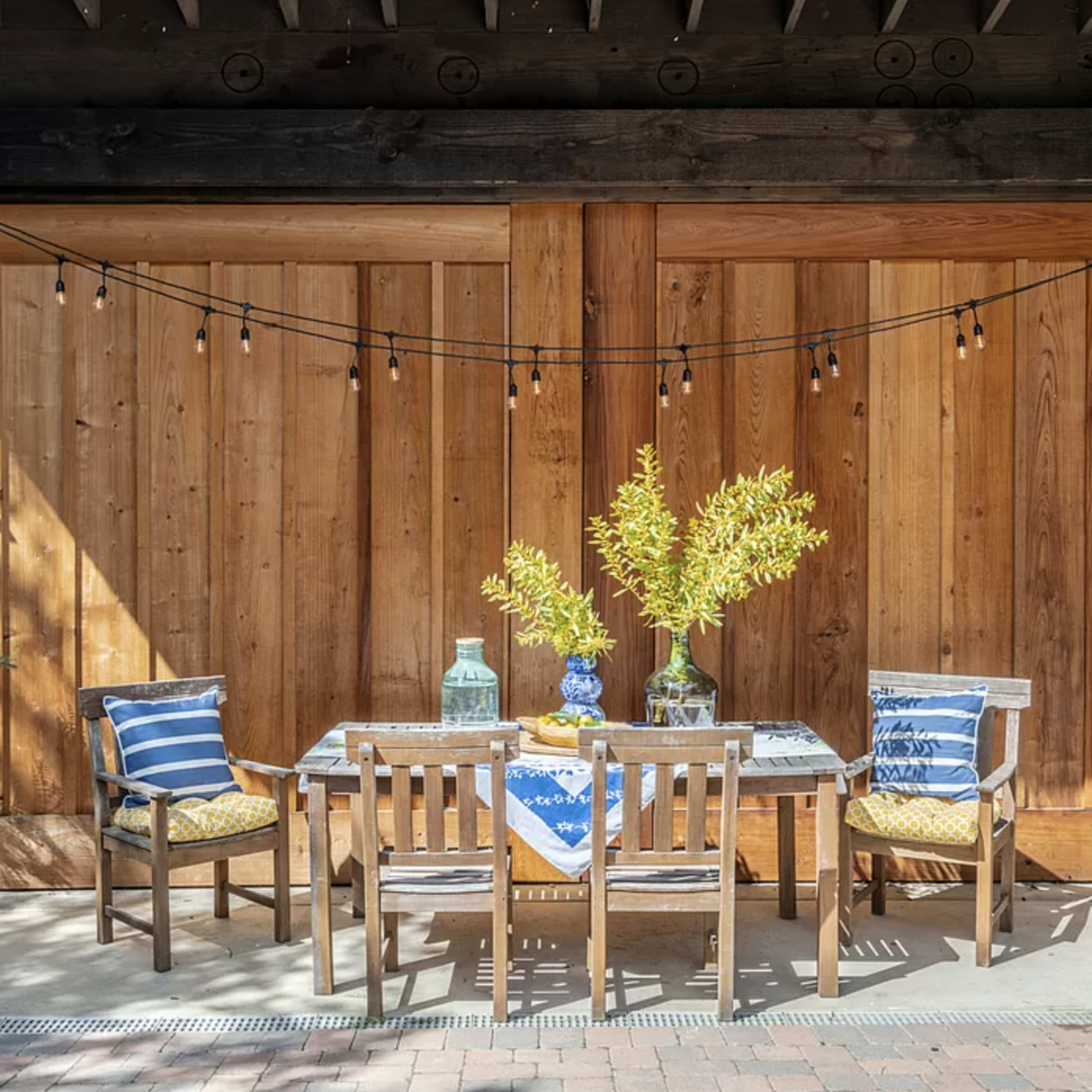 A wooden patio features a table set with a blue and white tablecloth, surrounded by three chairs and decorated with a vase of greenery.
