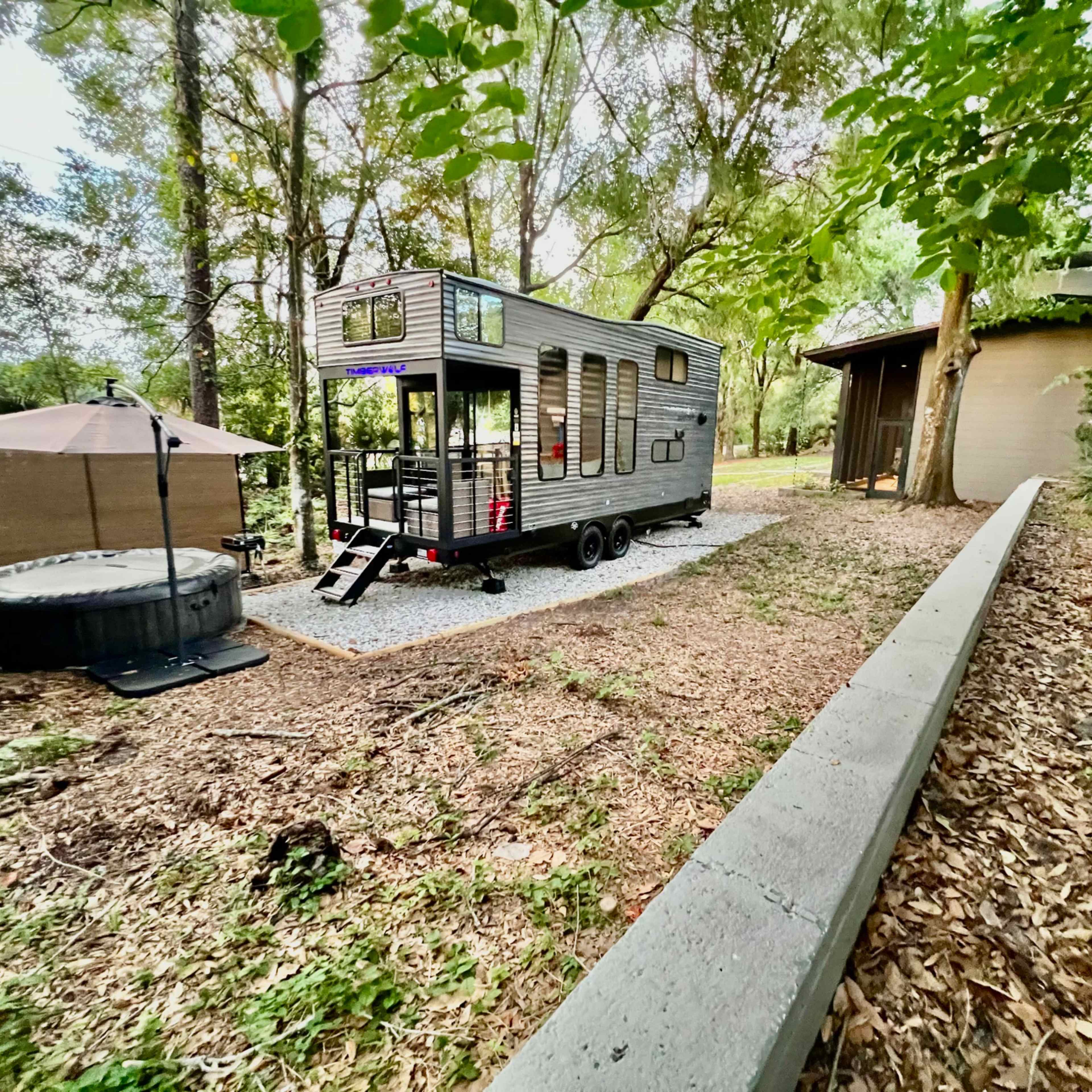 A tiny house on wheels is parked next to a circular building, surrounded by trees and a gravel path.