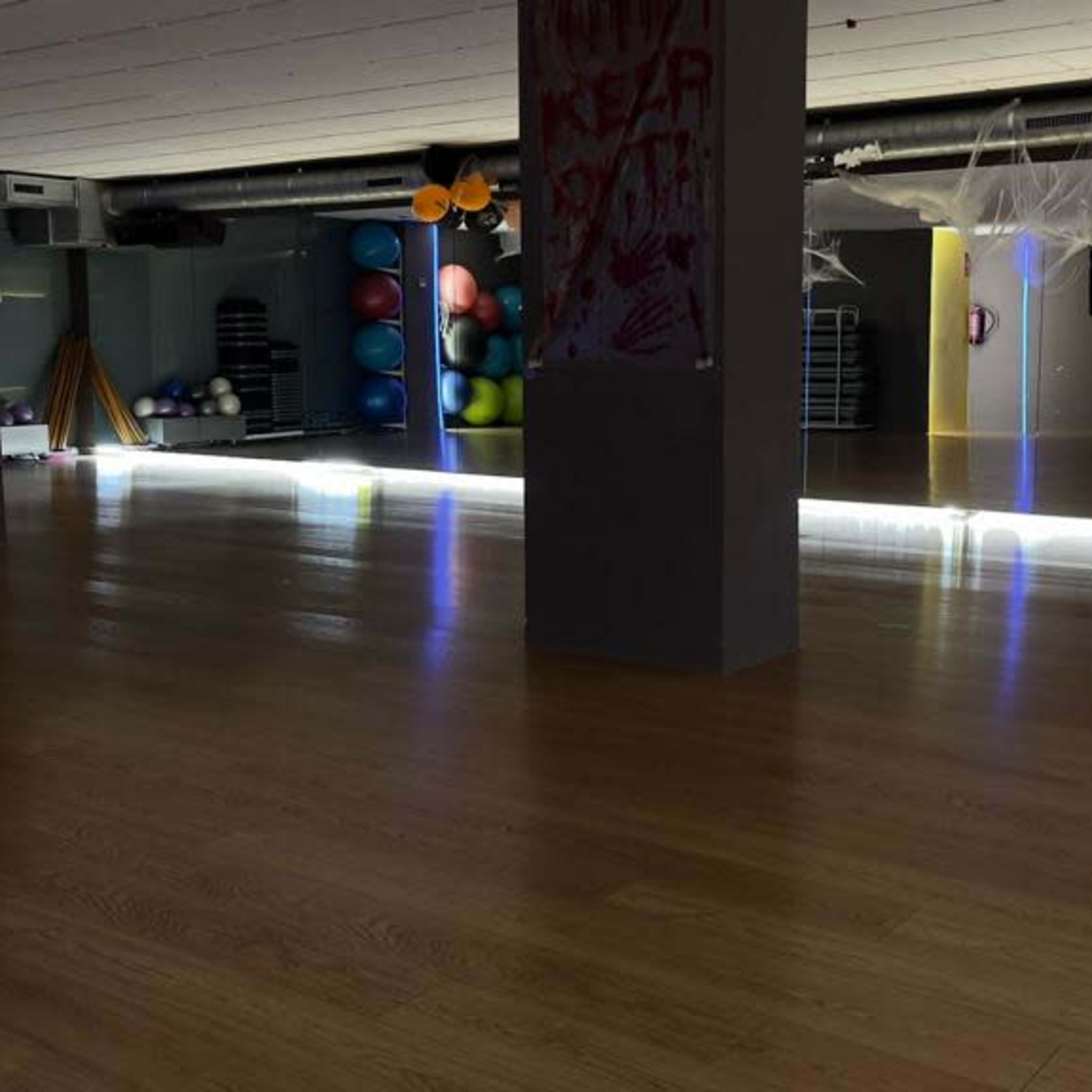 An empty gym studio with wooden flooring, mirrored walls, and exercise equipment along the sides, illuminated by lights.