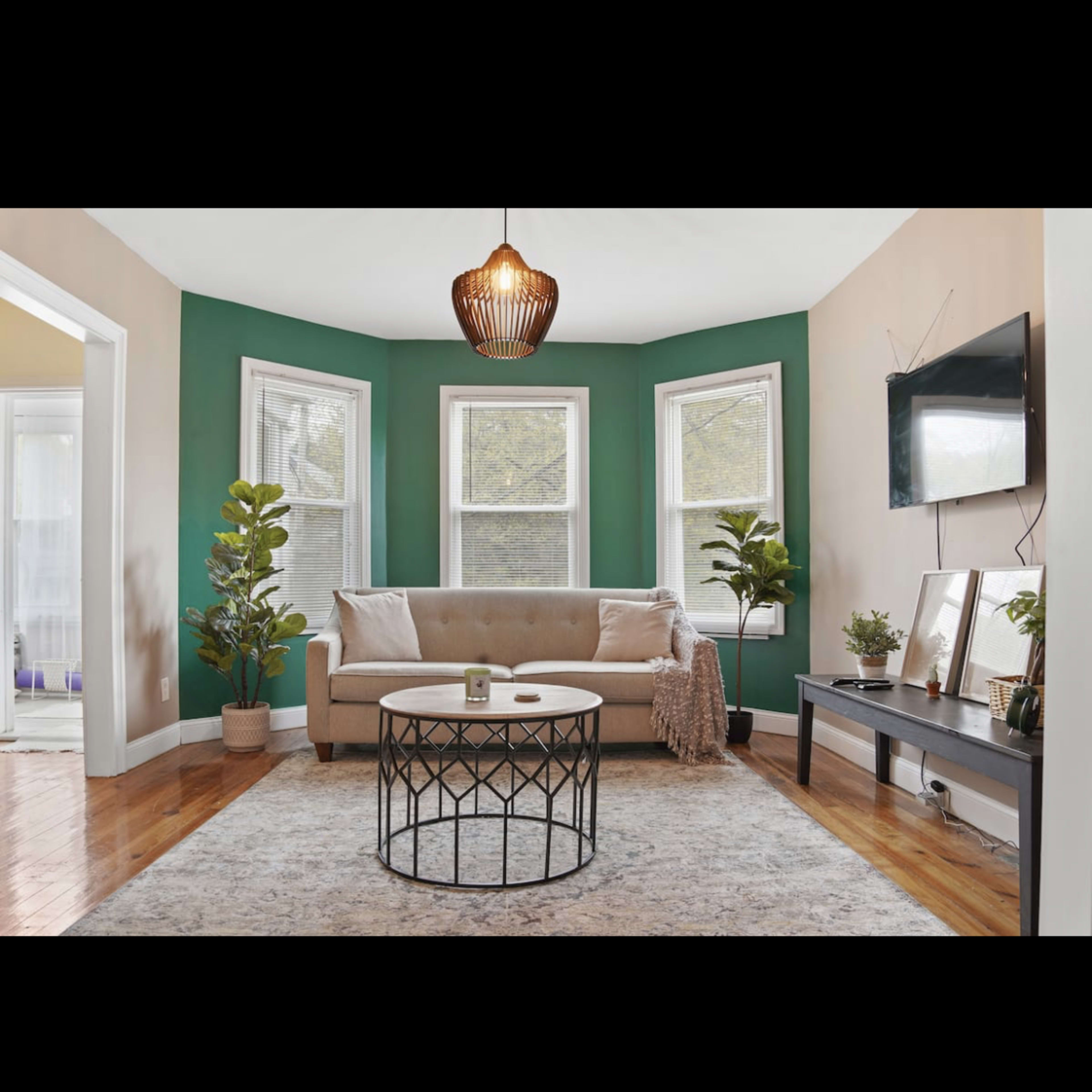 The living room features a beige couch, a round coffee table, green accent walls, and large windows that allow natural light to fill the space.