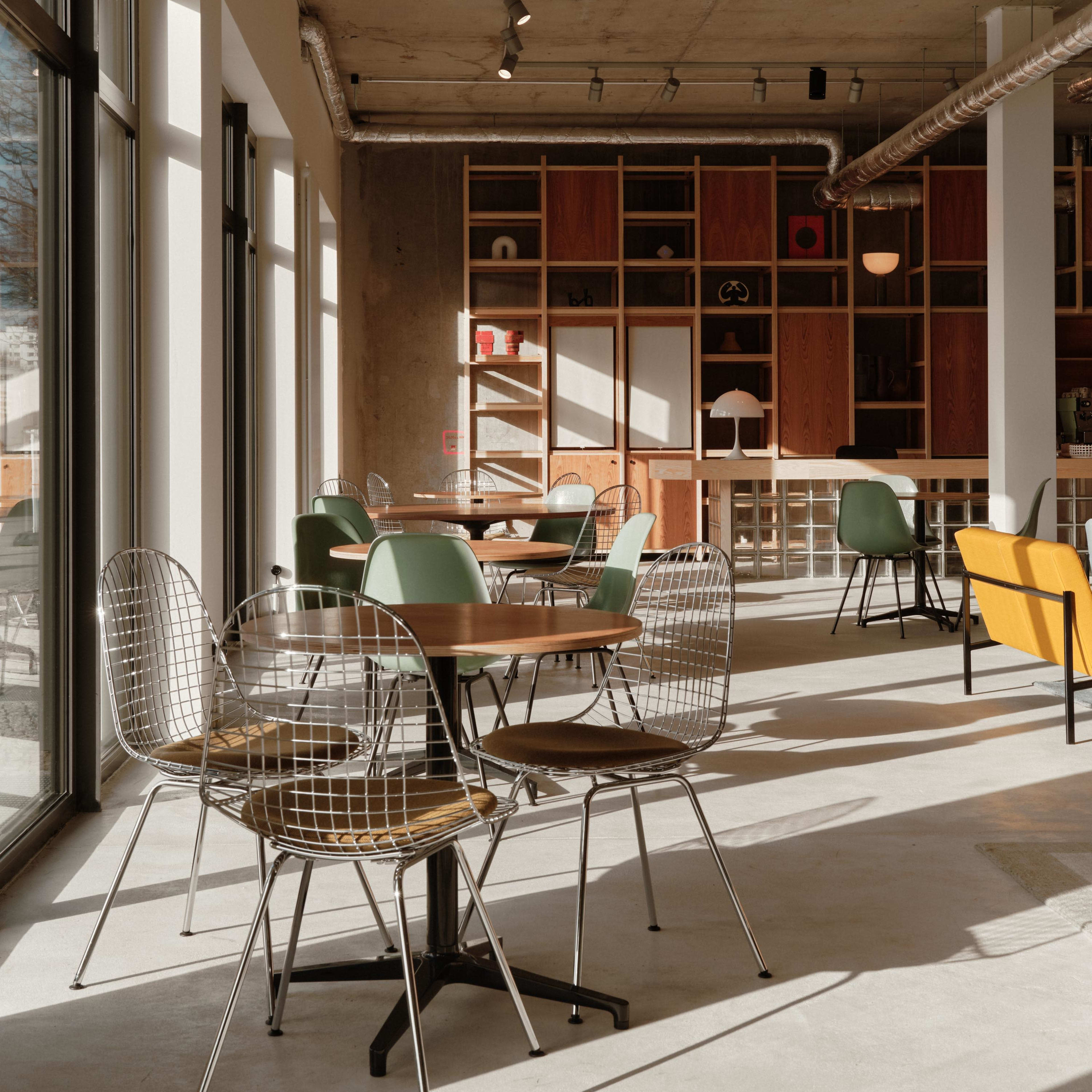 A modern café interior with various round tables and wire chairs, featuring a mix of colorful seating and a wooden shelving unit in the background.