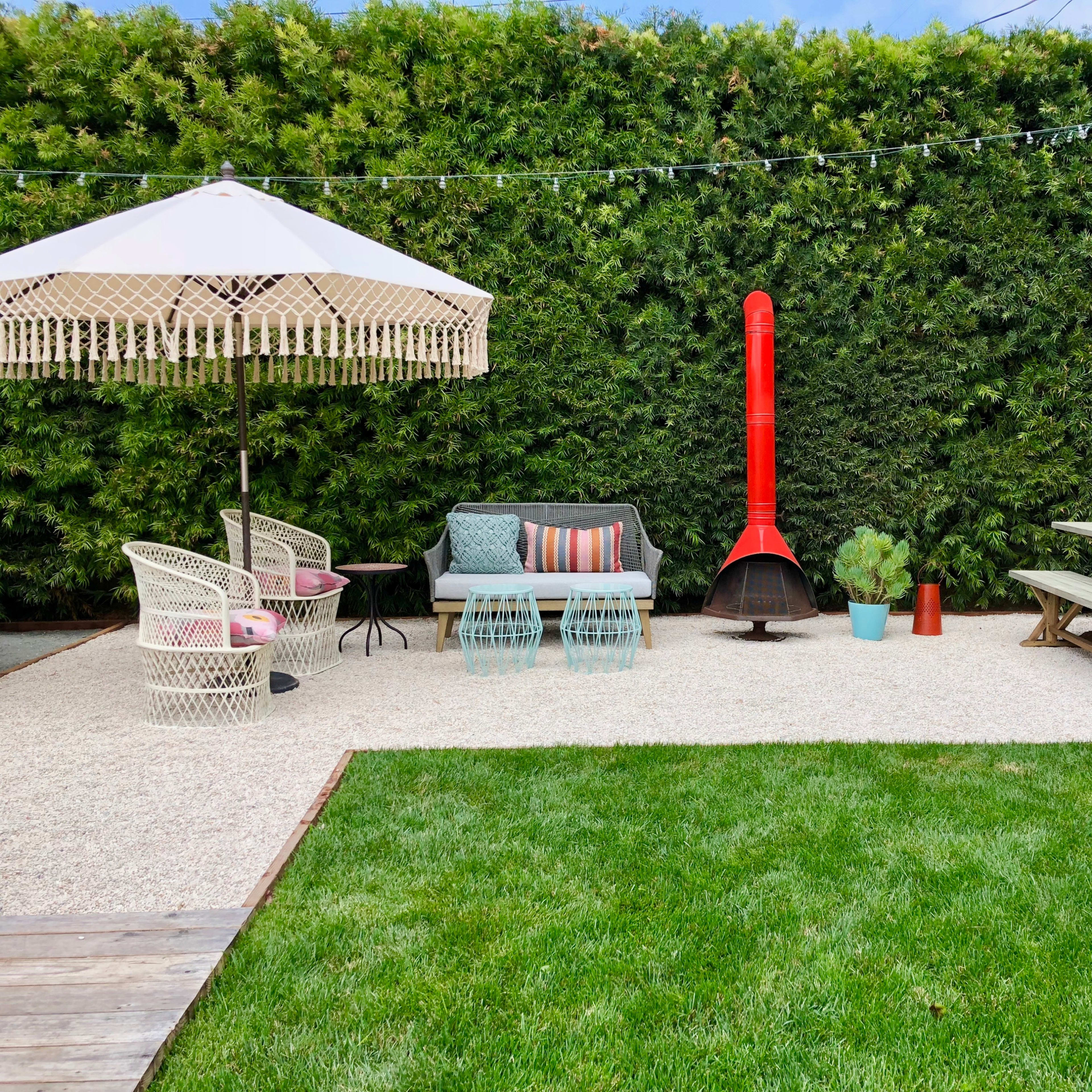 The outdoor space features a vibrant seating area with a red outdoor fireplace, surrounded by lush green hedges and a gravel patio.