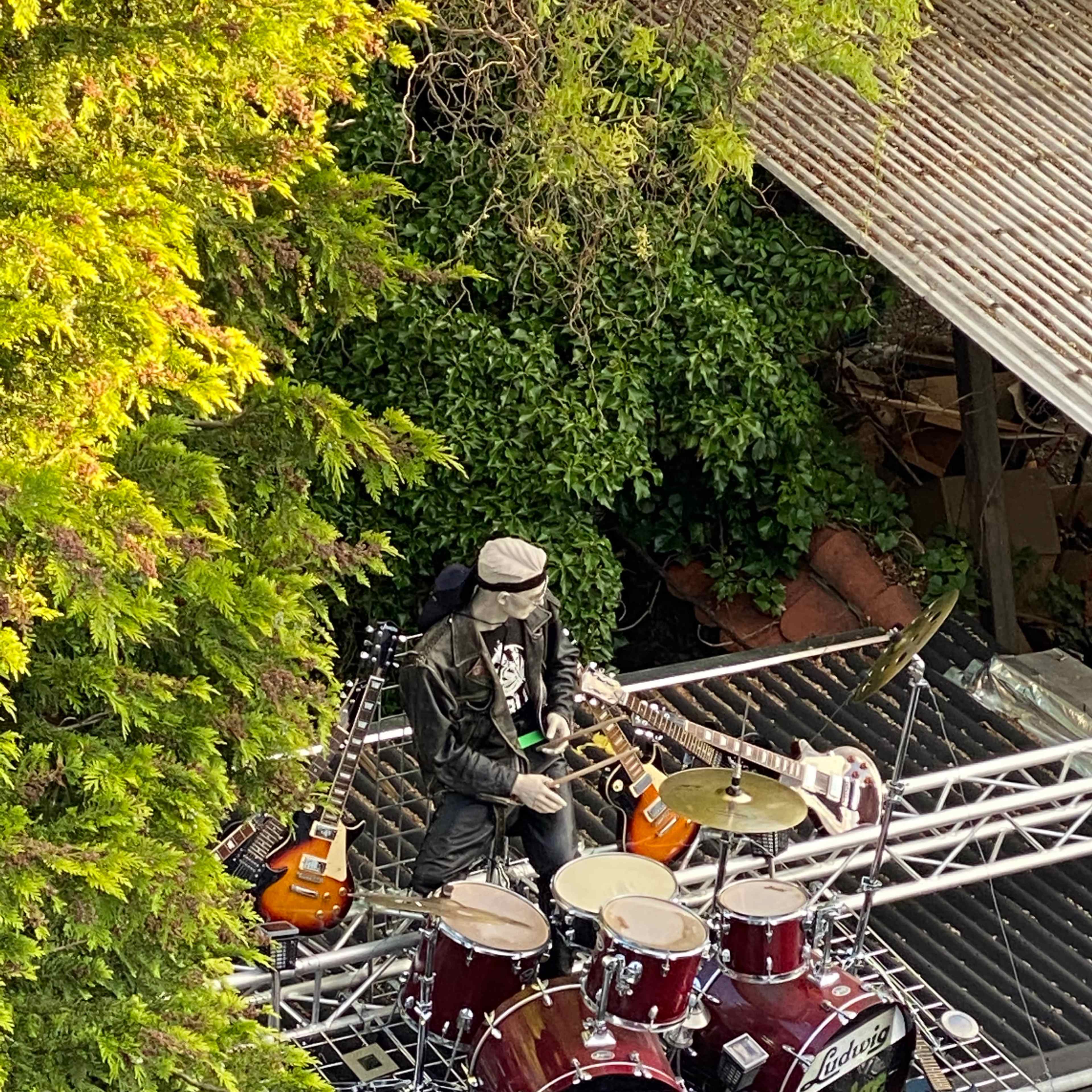 A drummer is playing a drum set positioned on a rooftop, surrounded by greenery and various musical instruments.