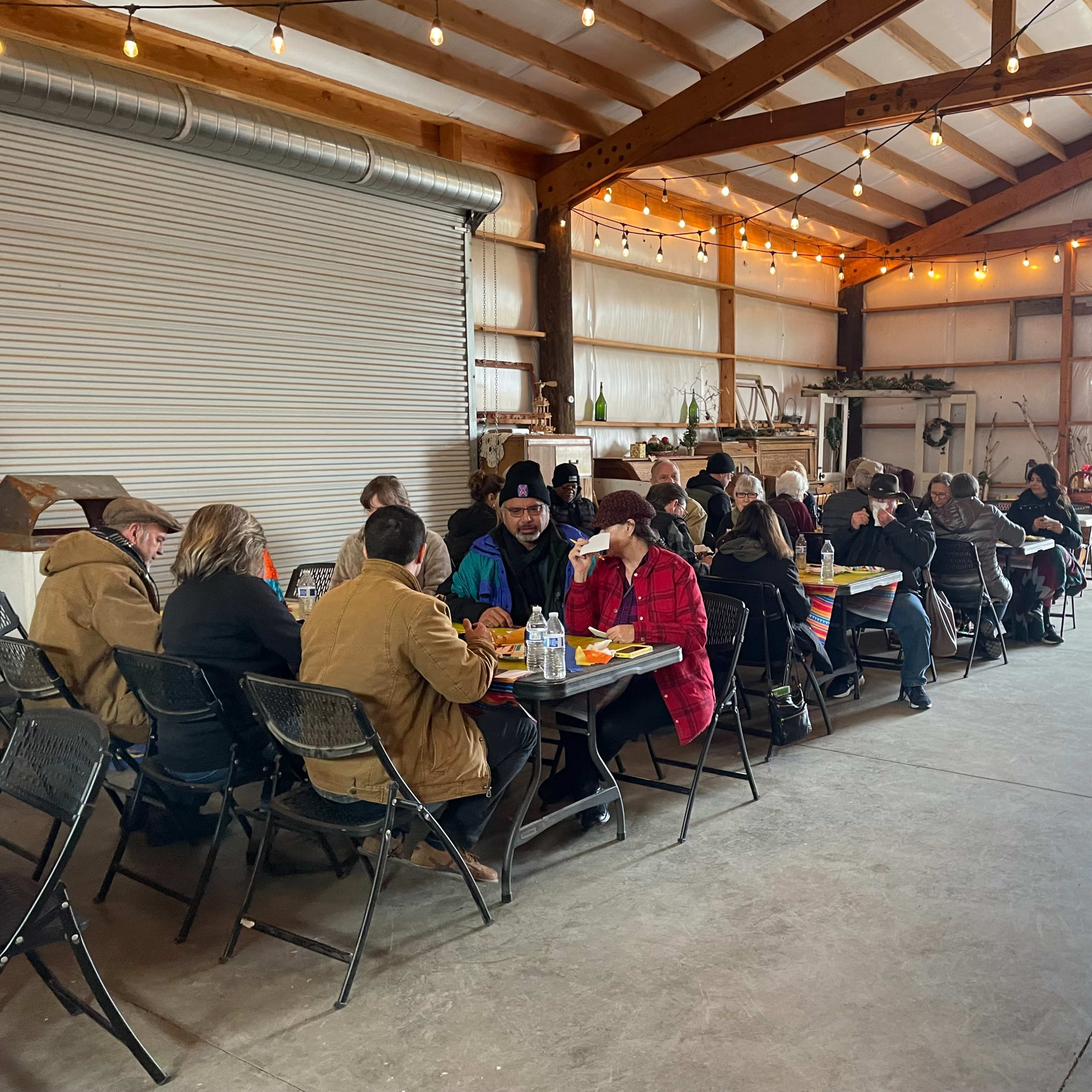 A group of people sits at tables in a spacious, well-lit barn, sharing a meal together.