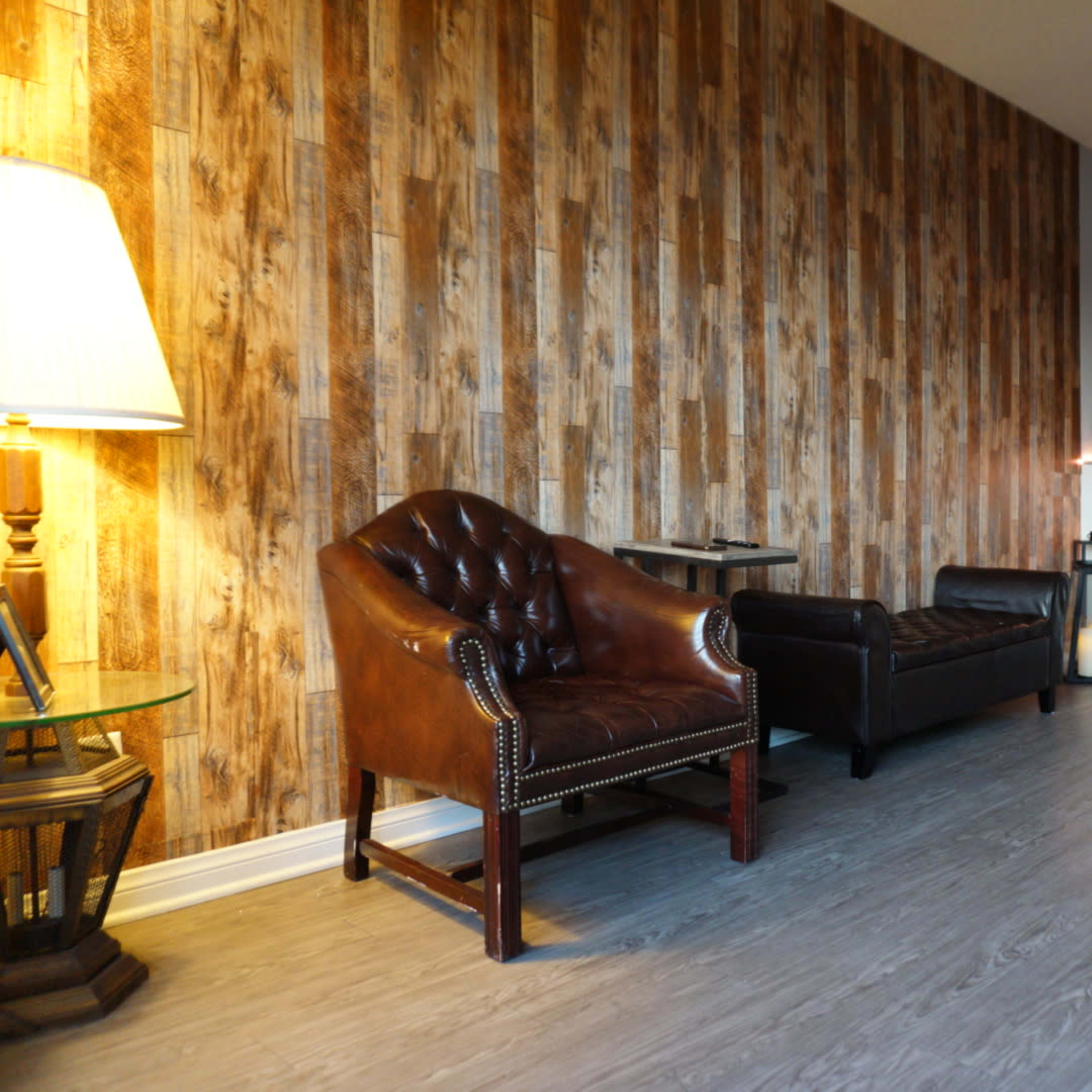A living room with a wood-paneled wall, a brown leather armchair, a dark leather sofa, a glass side table with a lamp, and a shelf with books and a small speaker.