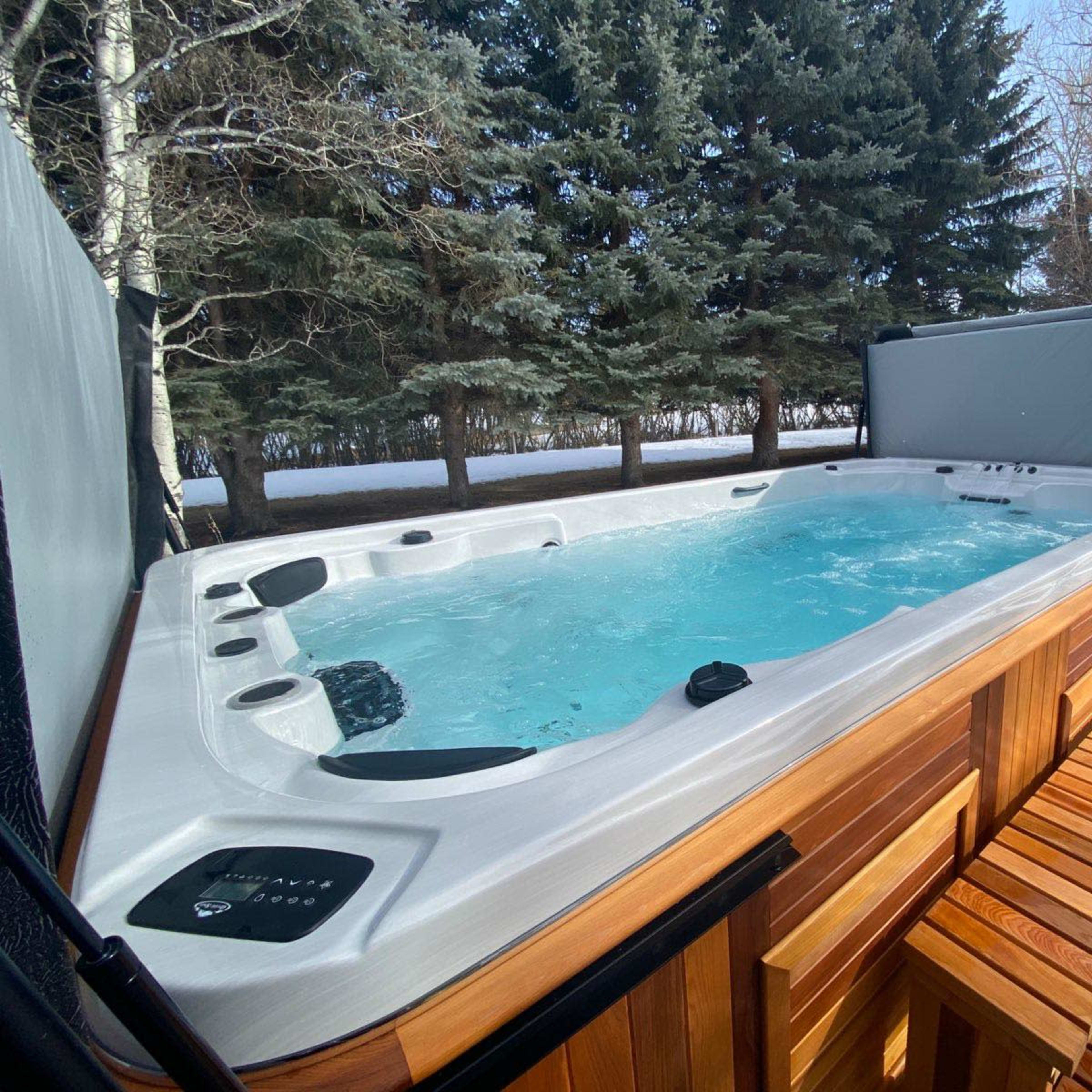 A rectangular hot tub with a wooden exterior and a covered top is situated outdoors near snow-covered ground and tall evergreen trees.
