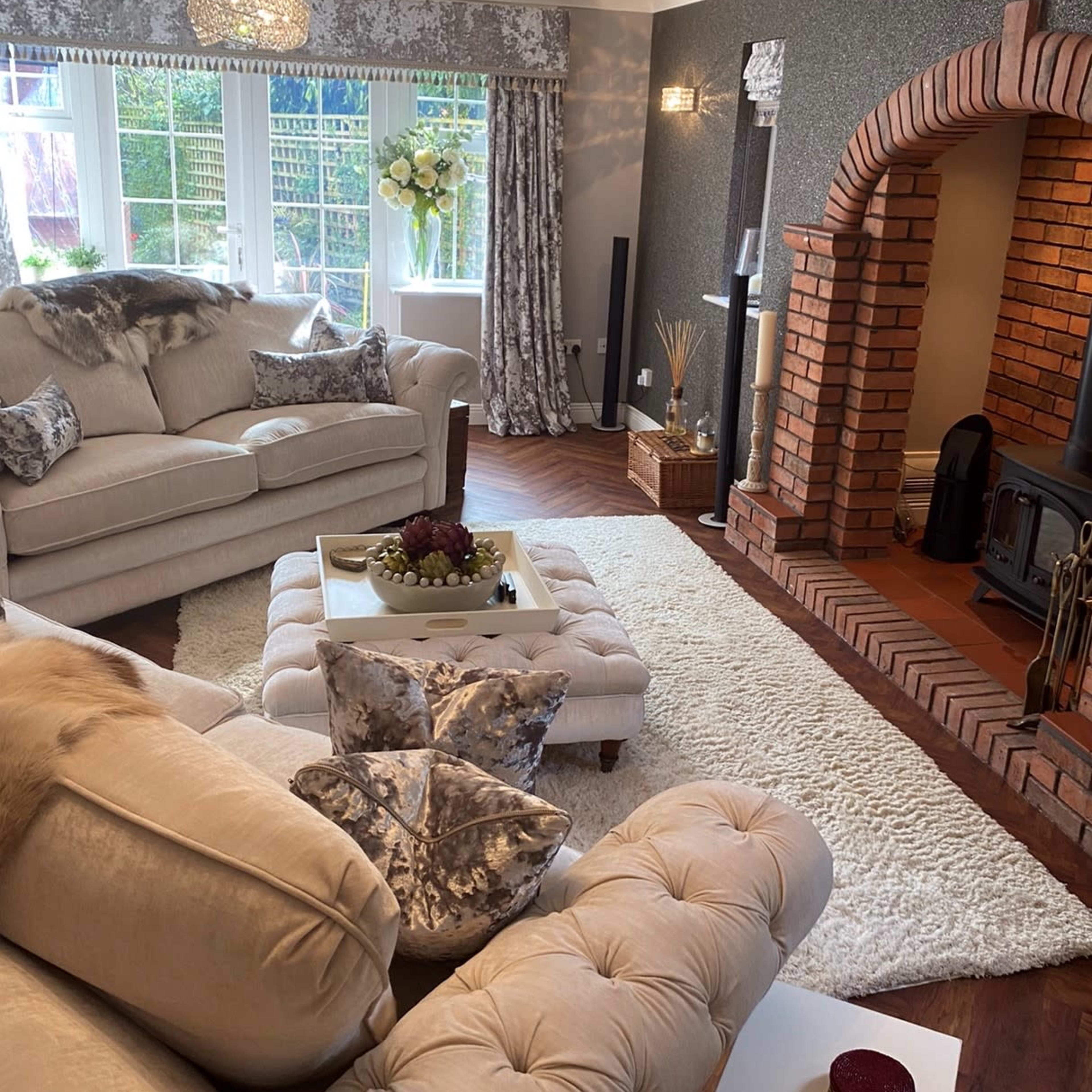 A cozy living room featuring a beige sectional sofa, a coffee table with decorative items, a plush area rug, and a brick fireplace with a stove.