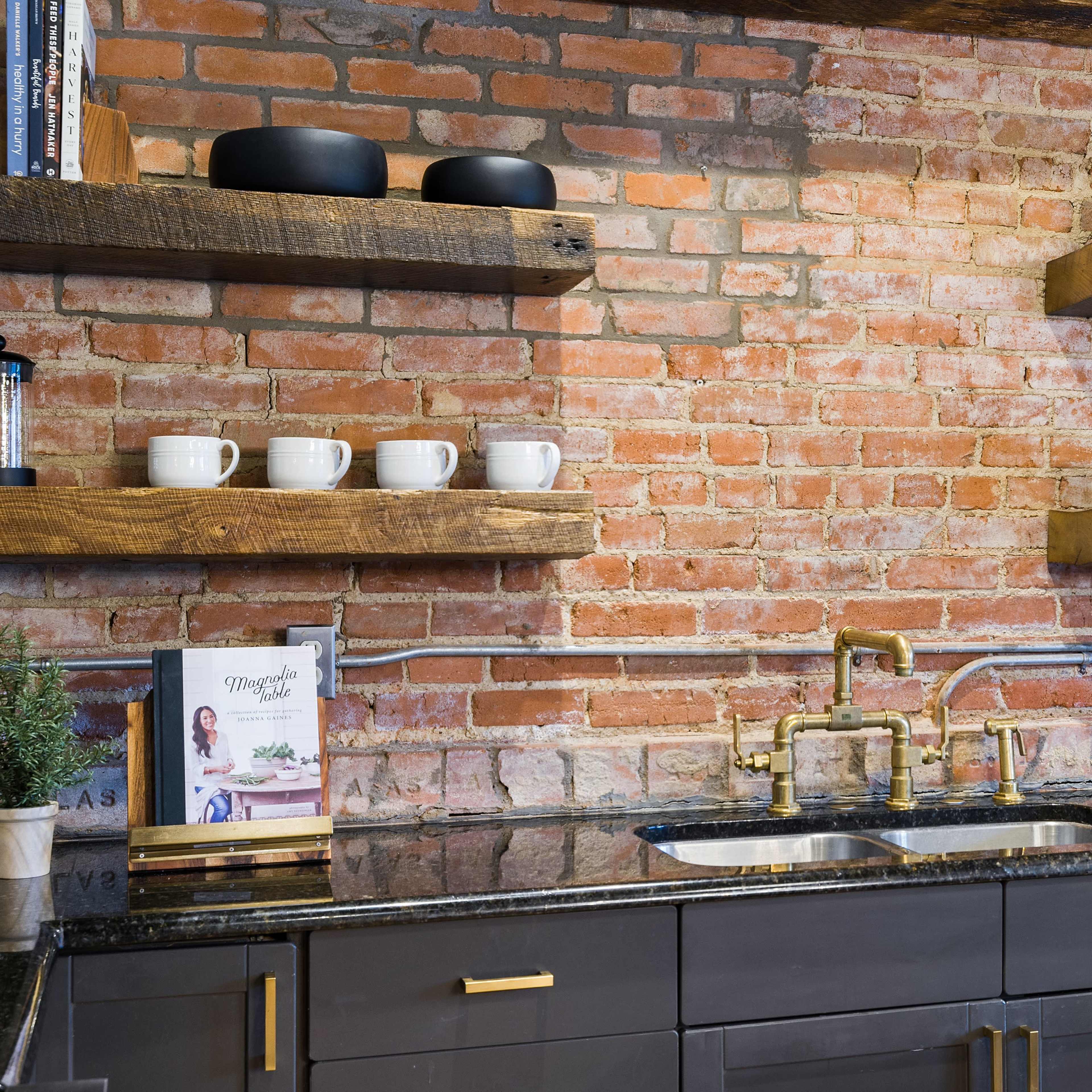 A kitchen with exposed brick walls, wooden shelves displaying dishware and decor, and a dark countertop with a sink and faucet.
