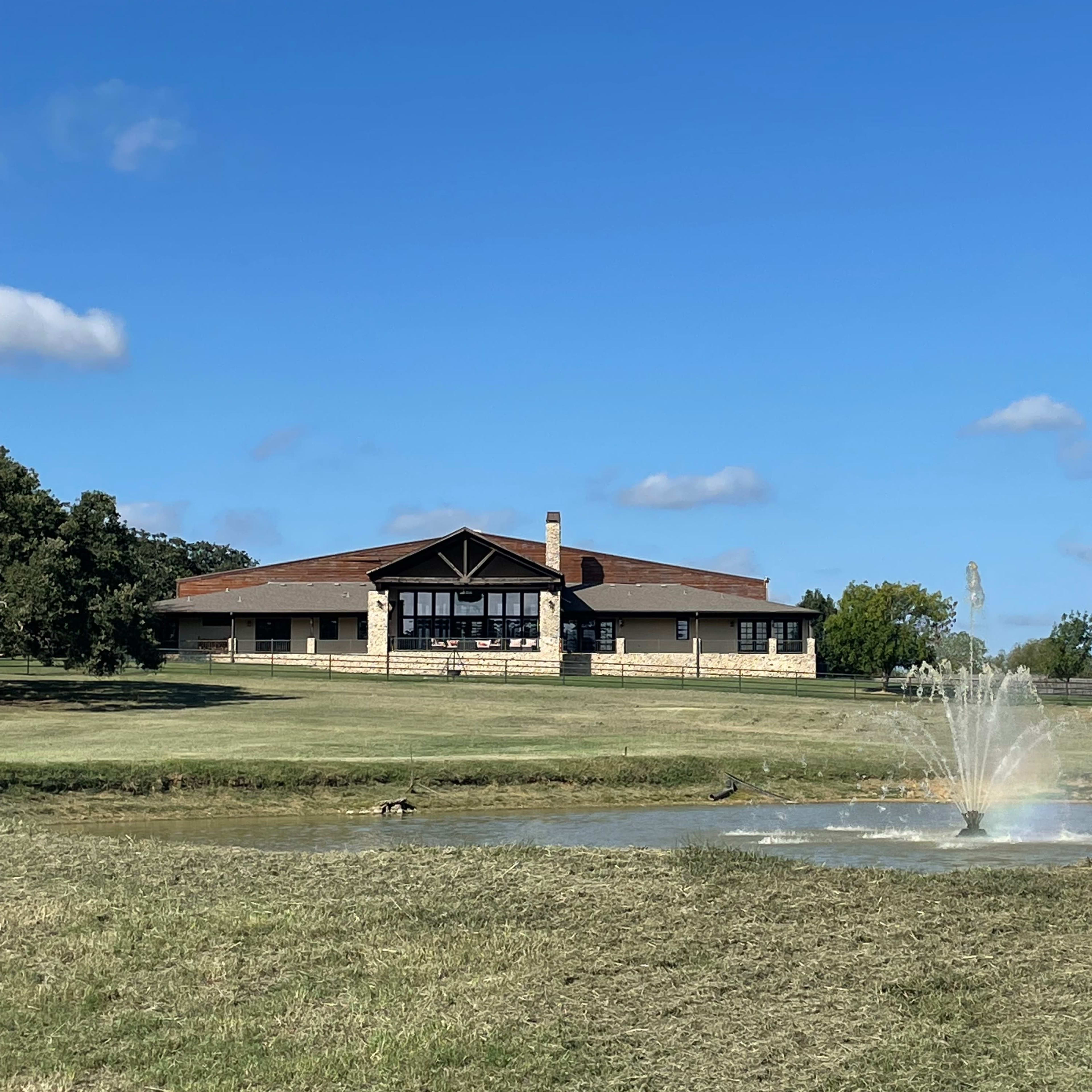 A large, modern building with a stone facade overlooks a pond featuring a fountain in a grassy area under a clear blue sky.