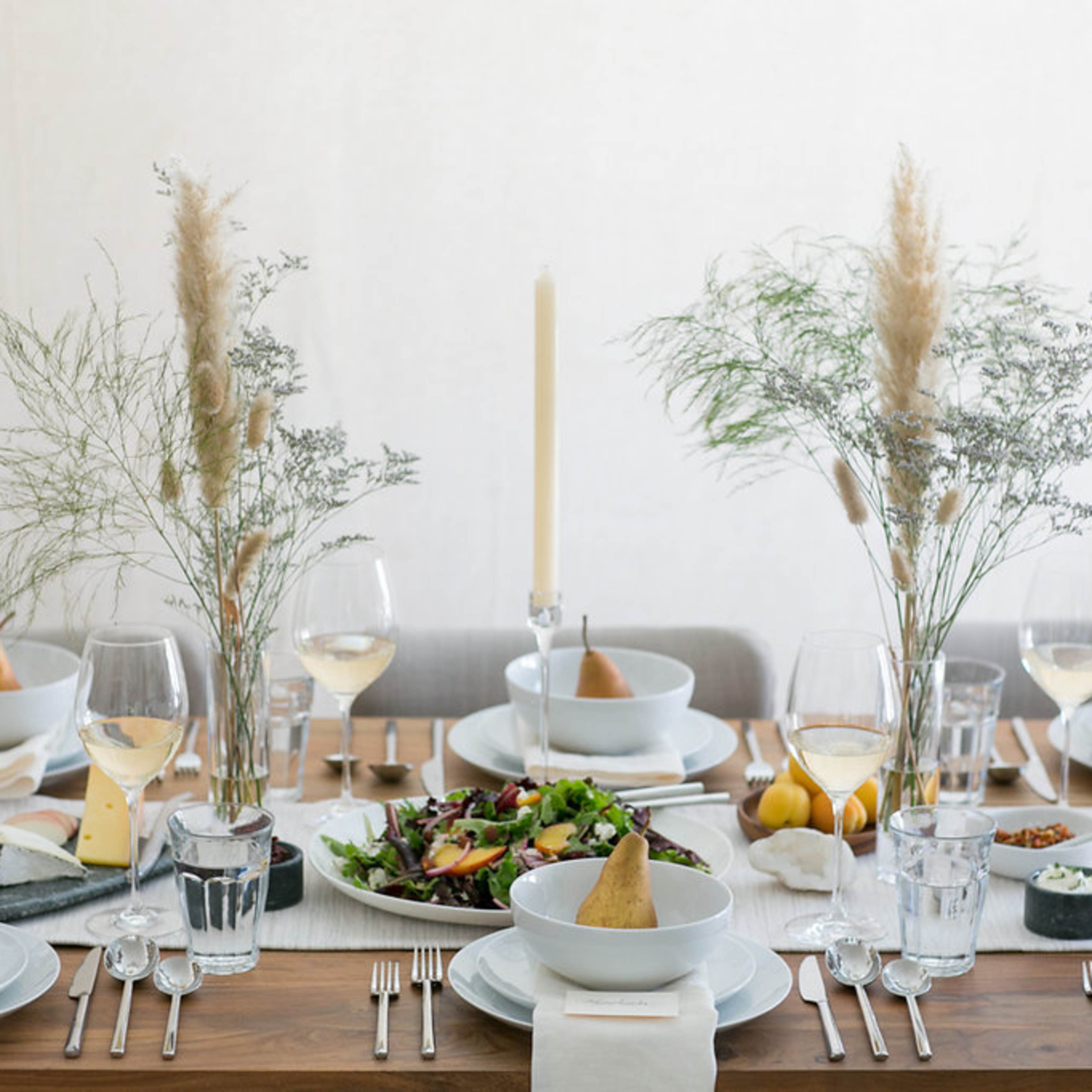 A long wooden table is set for a meal, featuring bowls of salad, glasses of wine, and decorative centerpieces with candles and greenery.