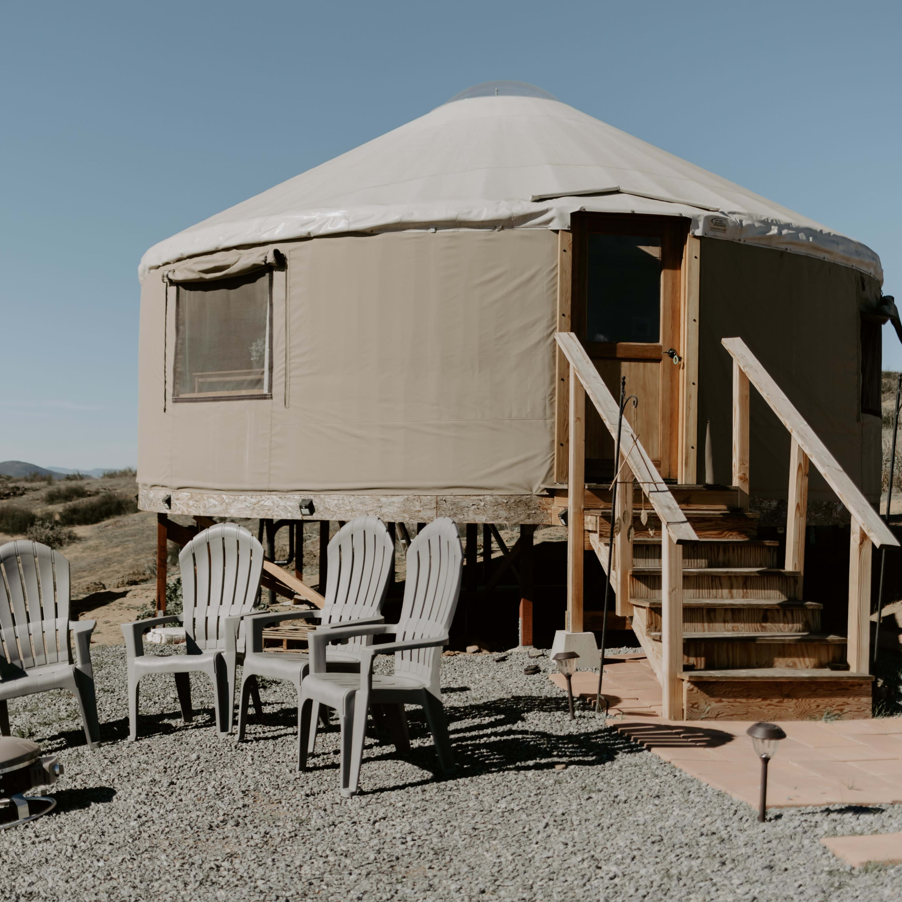 A yurt with a wooden deck and steps is surrounded by gravel and several plastic chairs.