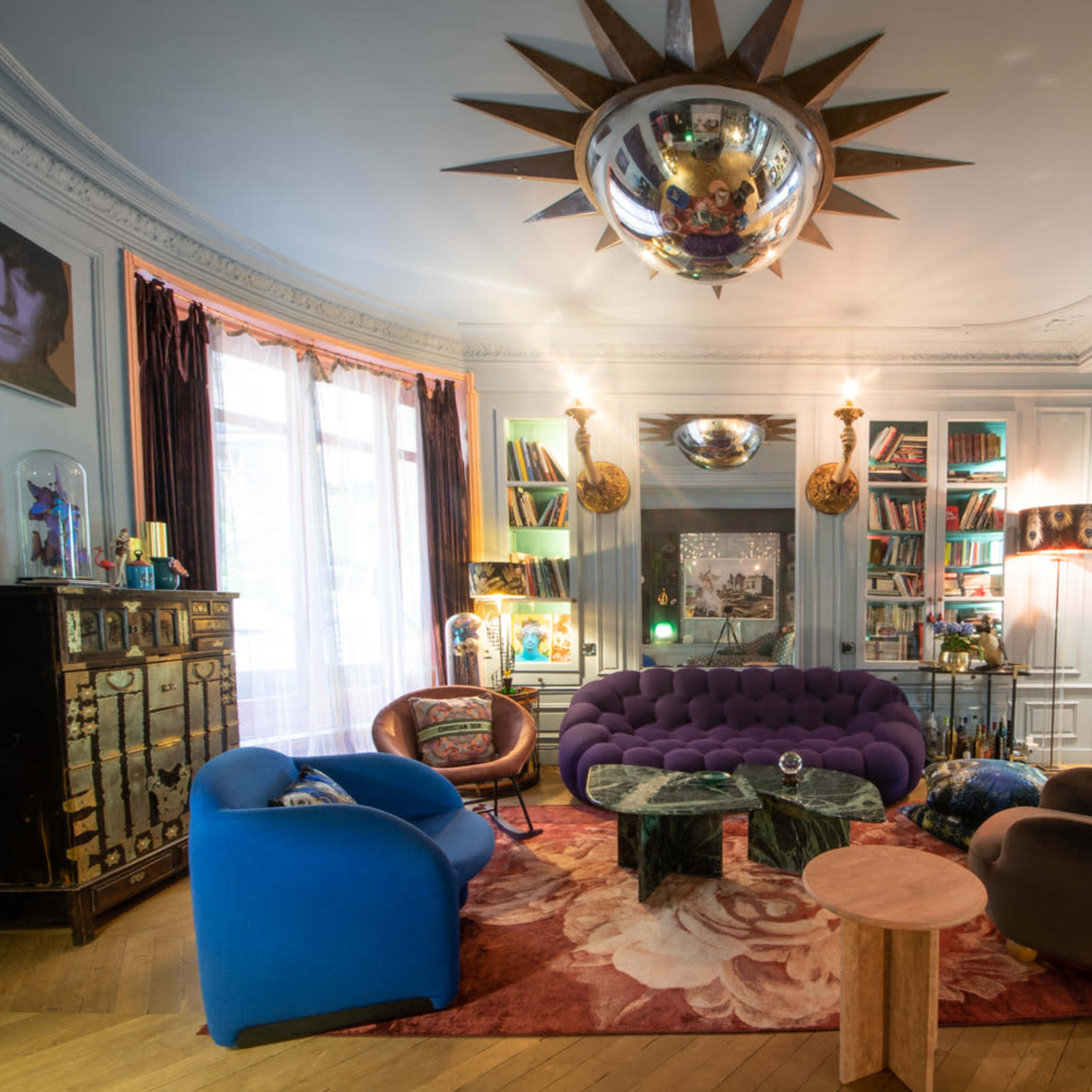 A stylish living room featuring a purple tufted sofa, two uniquely shaped armchairs, a round wooden coffee table, and a decorative ceiling fixture, all arranged around a floral area rug.