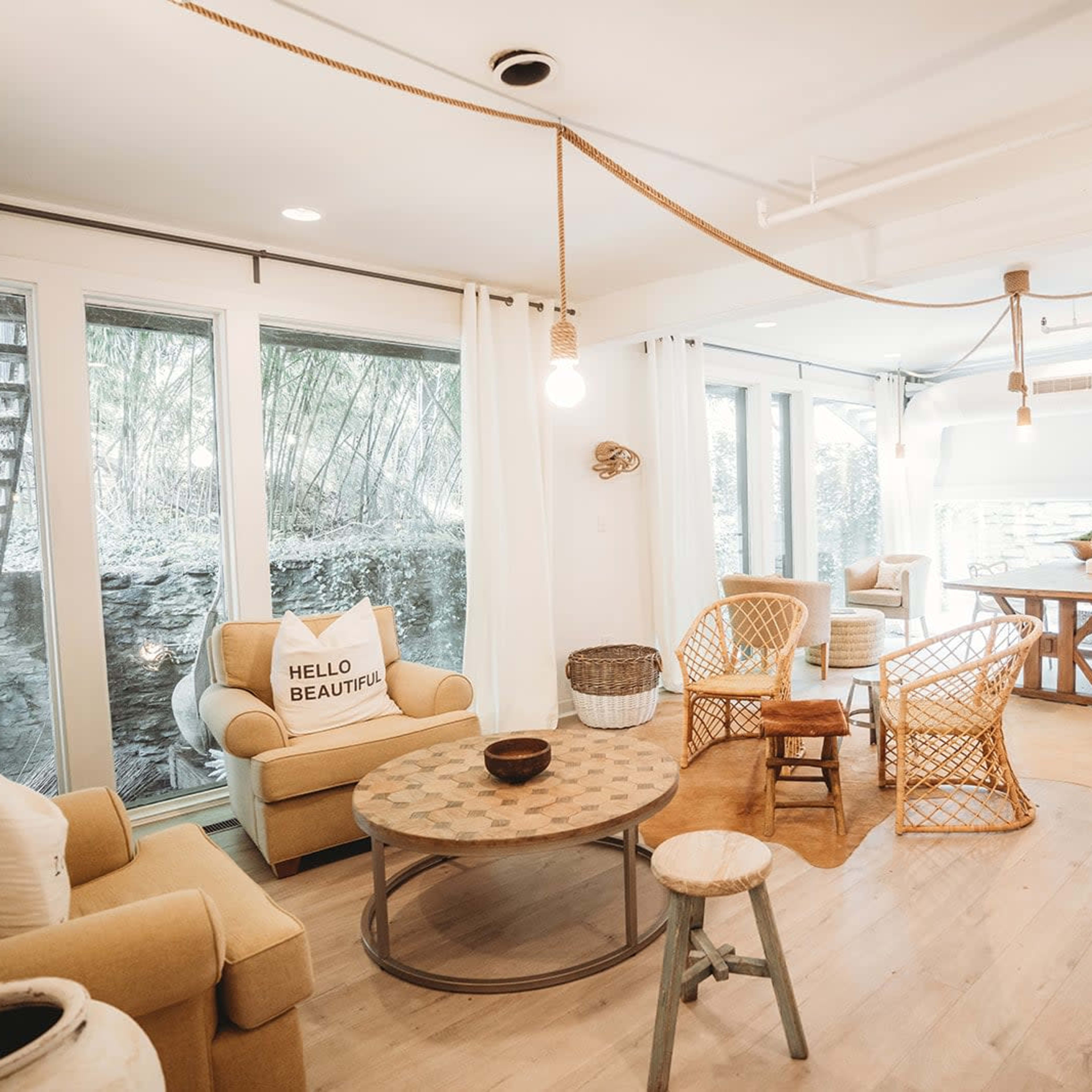 A cozy living space with a mix of wicker and upholstered furniture, a large table, and large windows allowing natural light to enter.
