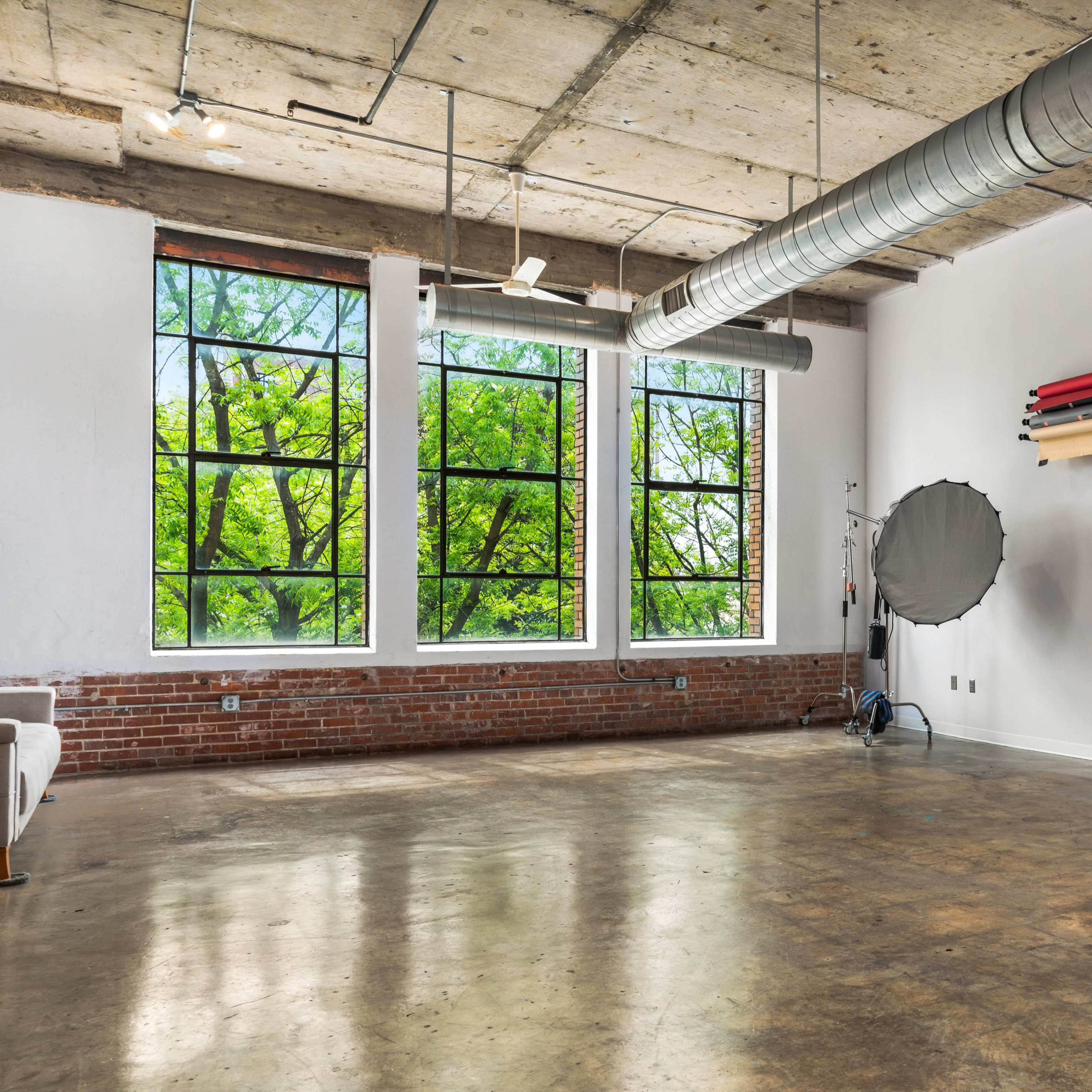 A spacious studio features large windows with views of green trees, exposed concrete ceilings, and photography equipment along the walls.