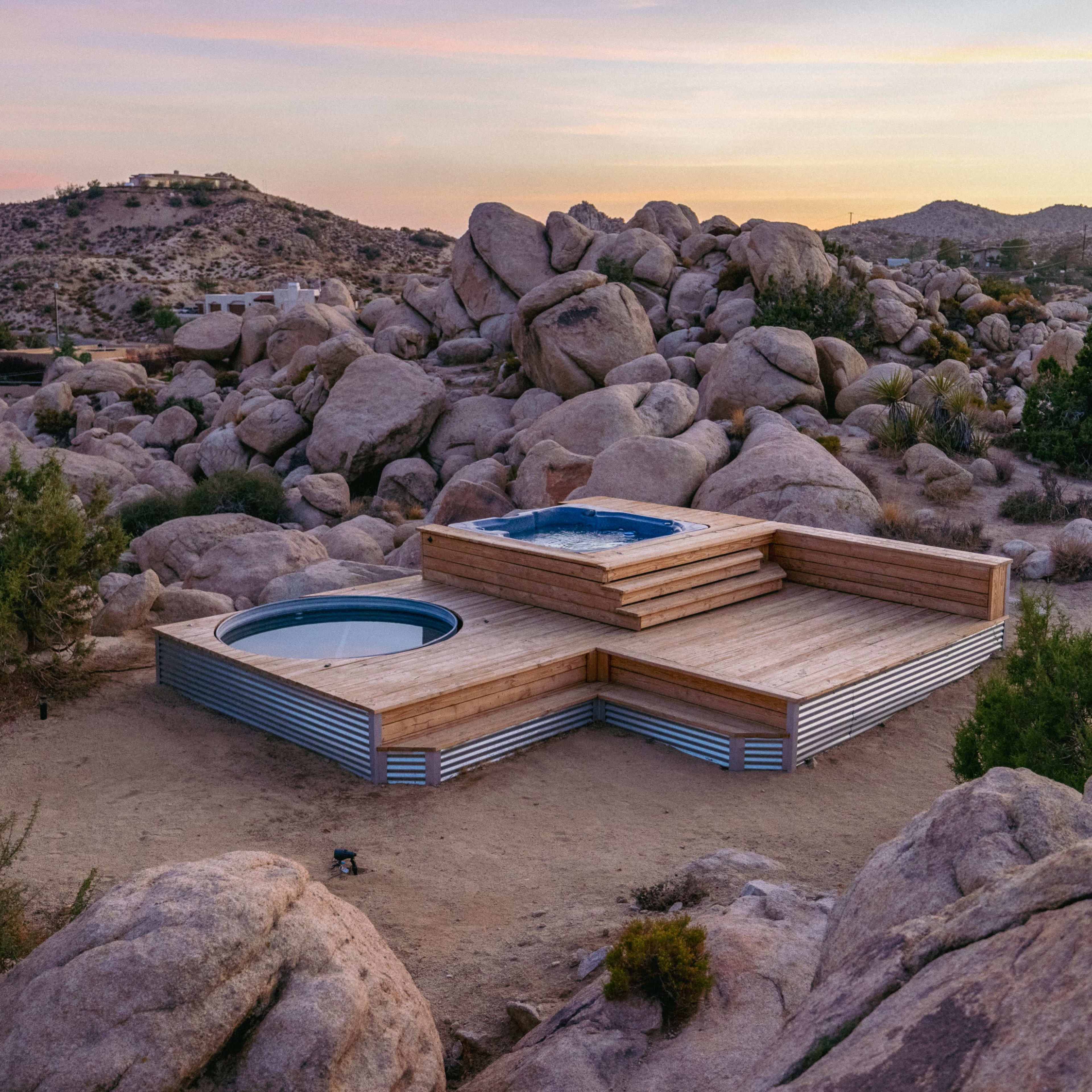 A wooden deck with a hot tub and a circular pool is nestled among large boulders in a desert landscape at sunset.