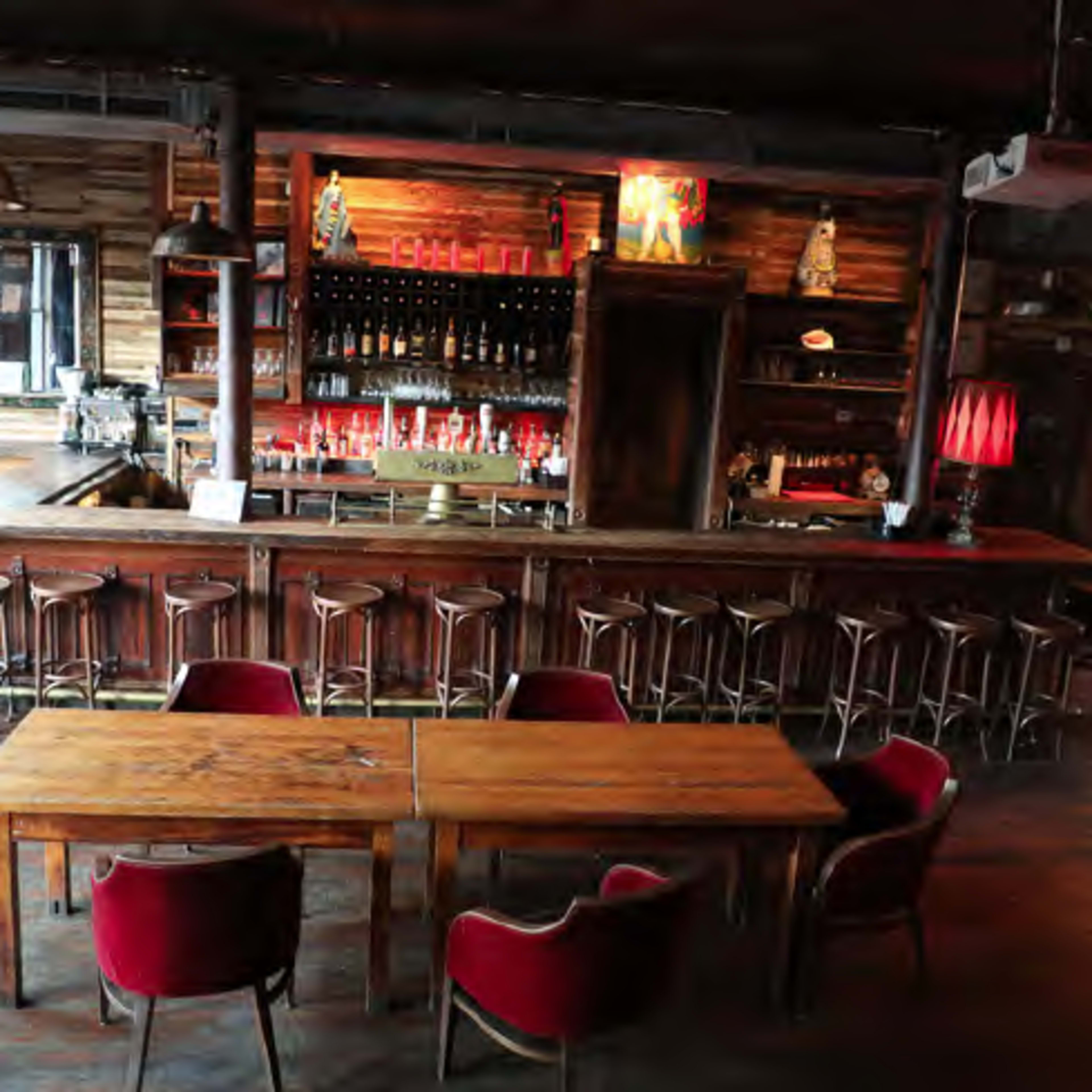 A dimly lit bar with wooden counters, shelves filled with bottles, and red velvet chairs arranged around wooden tables.