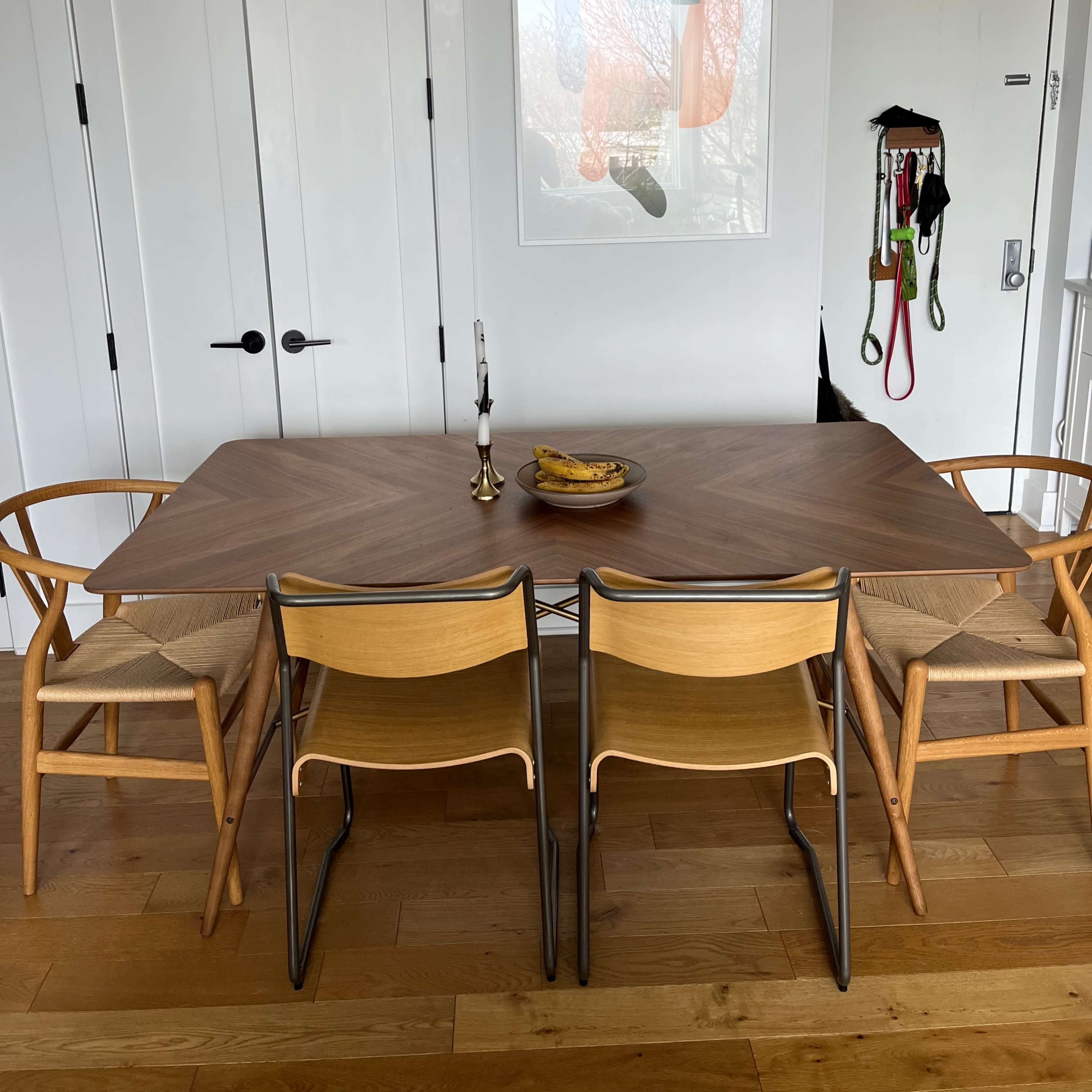 A dining table with wooden chairs and a candle holder is set against a wall featuring a framed artwork and an open door.