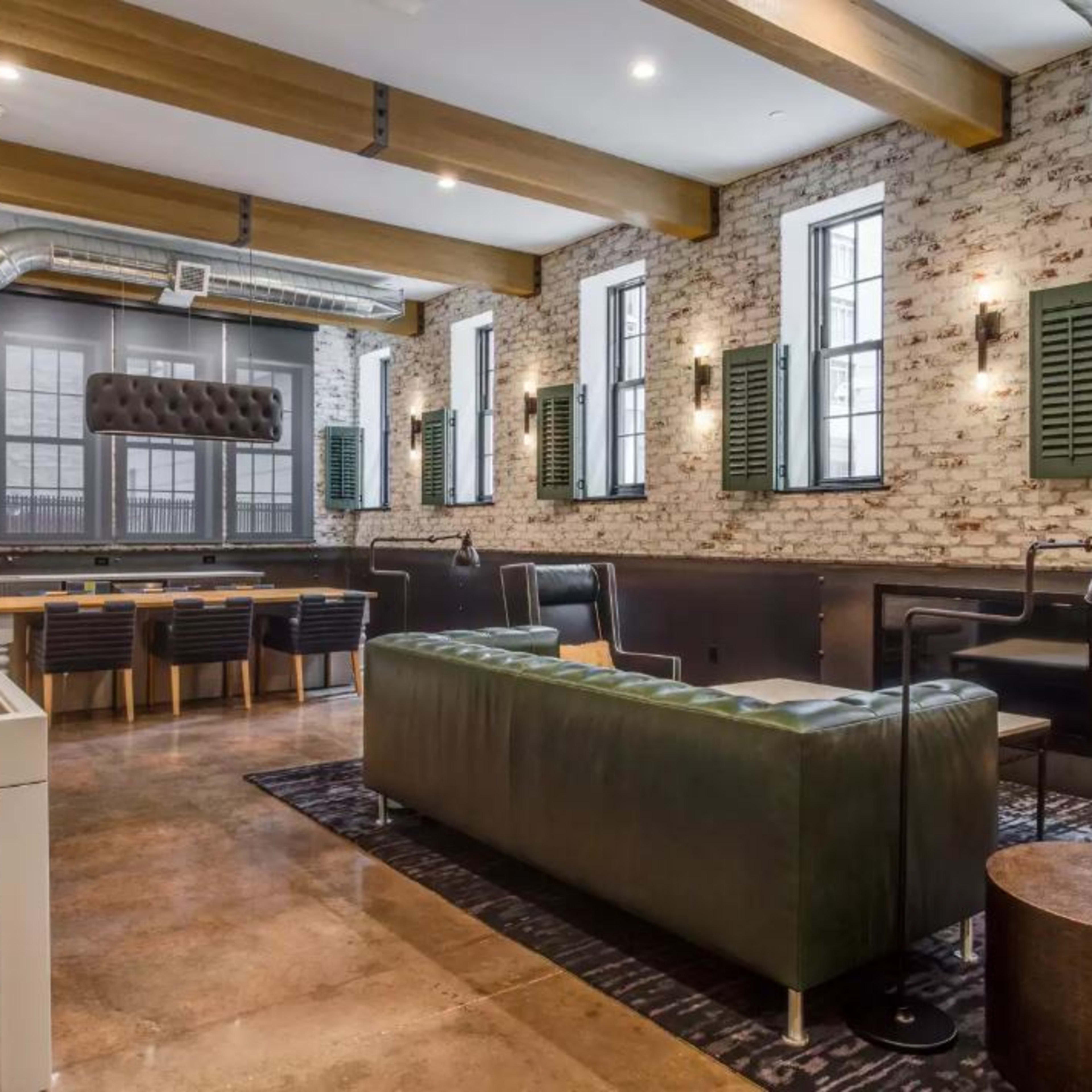 A spacious, industrial-style room with exposed brick walls, large windows, and a mix of seating arrangements including a green leather sofa and a wooden dining table.