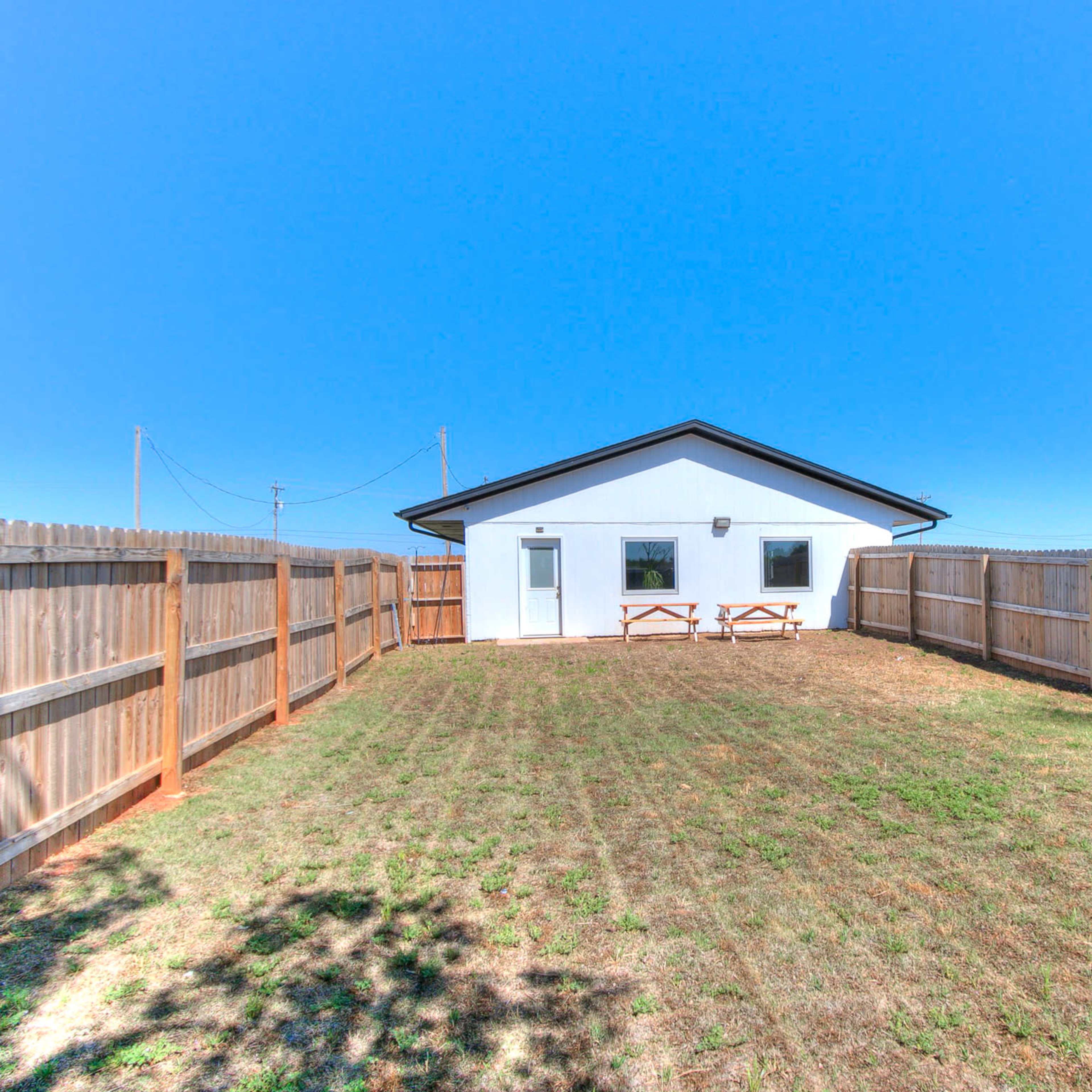 A fenced backyard features a grassy area and a simple building with two windows and a door.