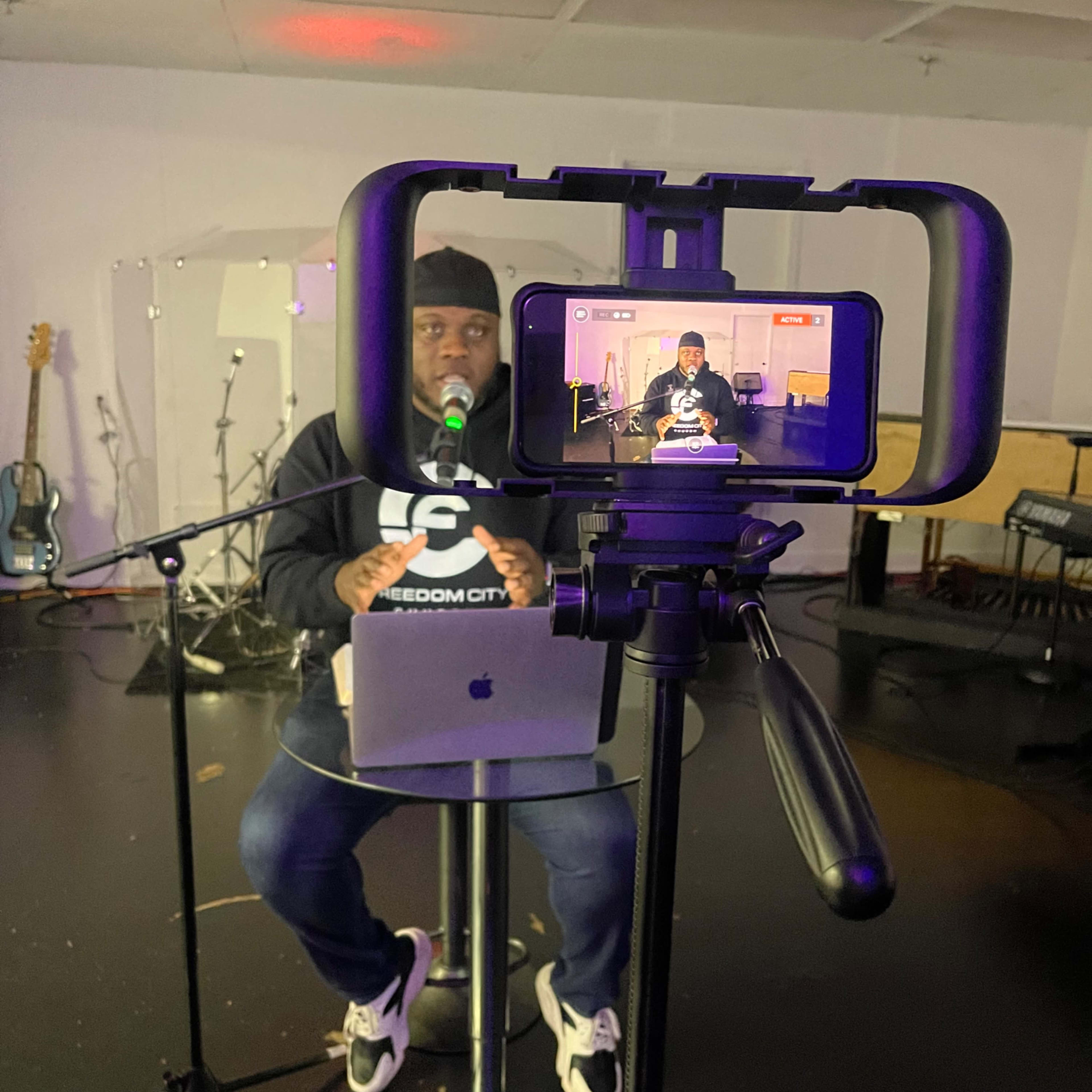 A person is speaking into a microphone while seated at a table, with a smartphone on a tripod capturing the scene.