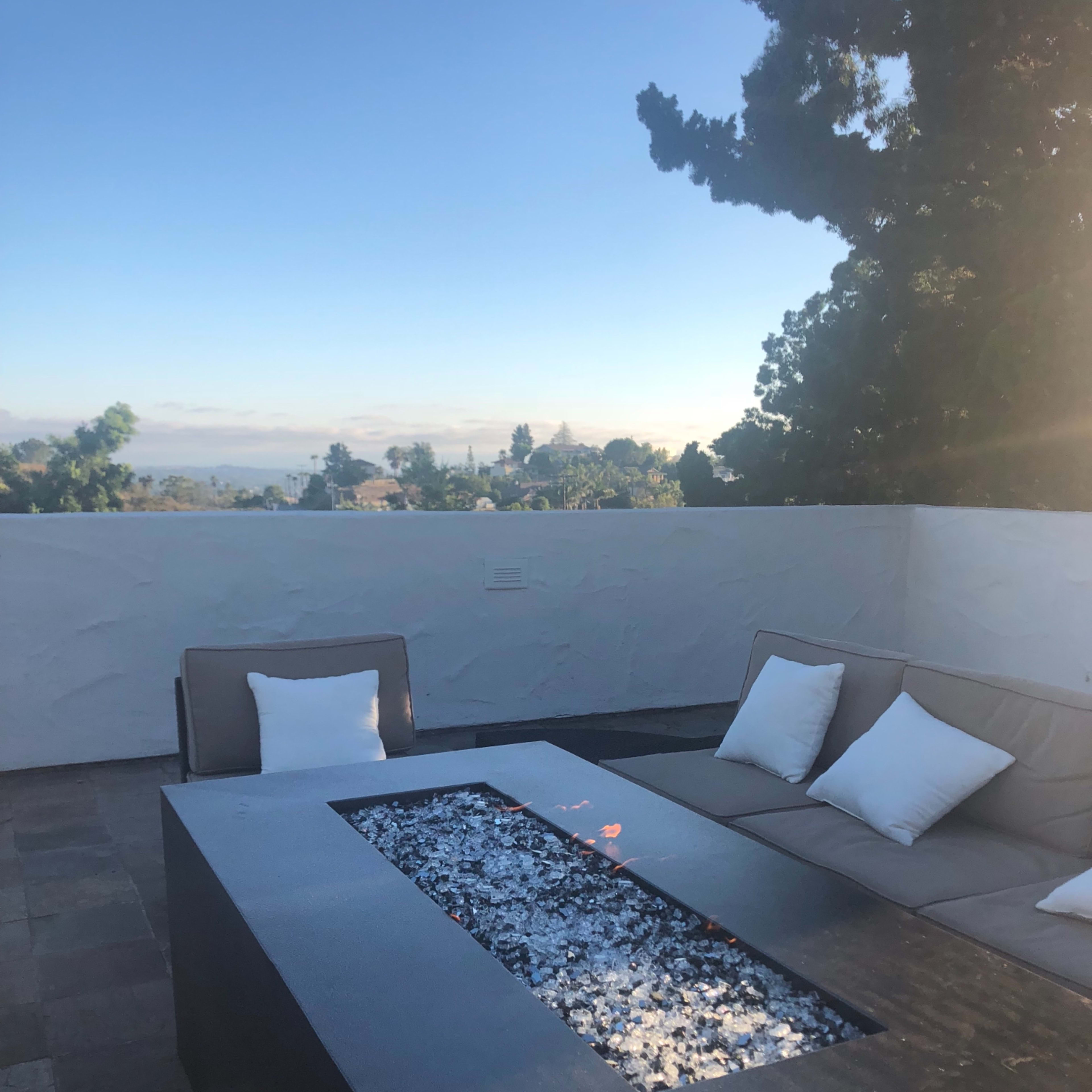 A modern outdoor seating area features a fire pit with a glass stone surface and a sectional sofa with white cushions, set against a clear sky and distant landscape.