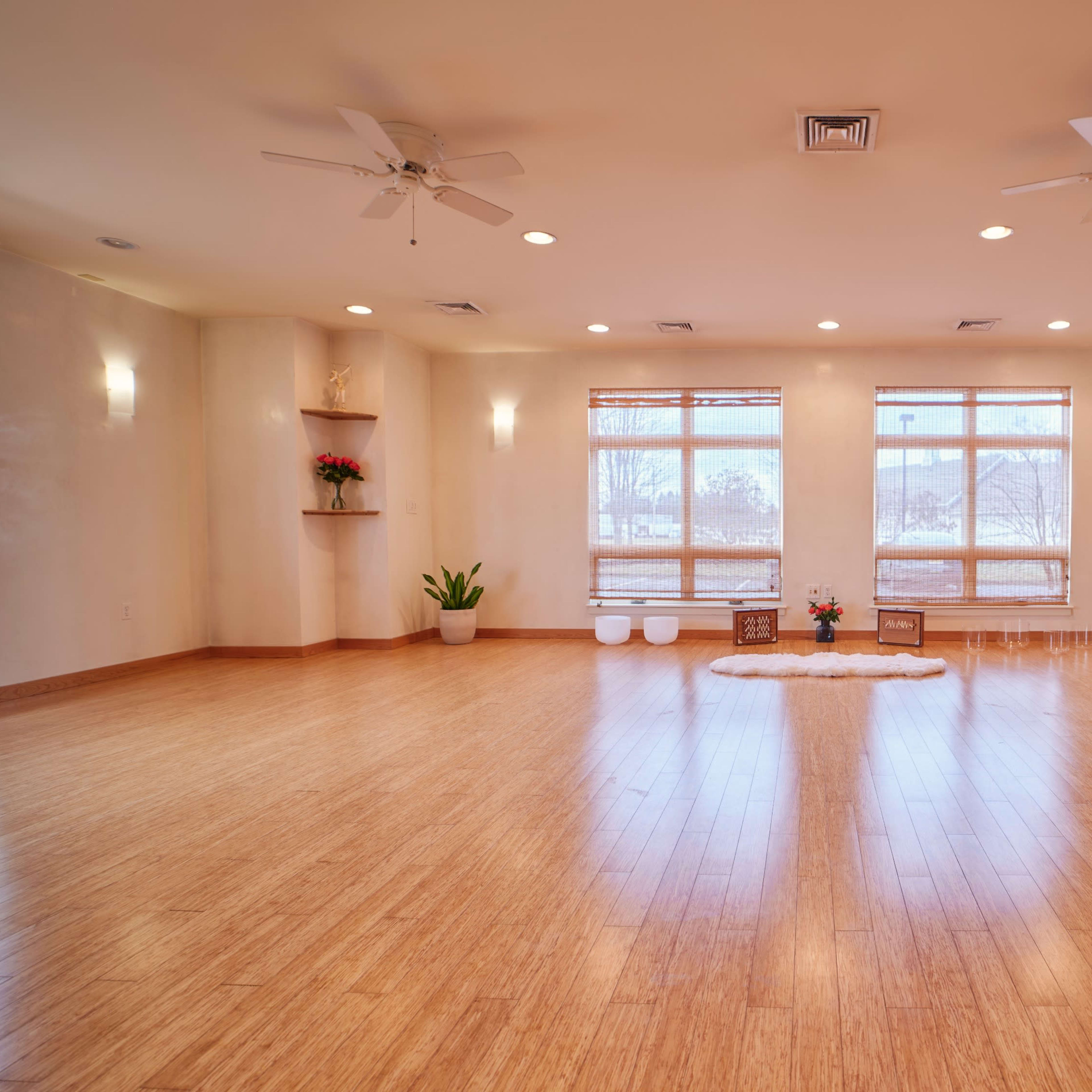 A spacious yoga studio with bamboo flooring, large windows, and several plants arranged around the room.