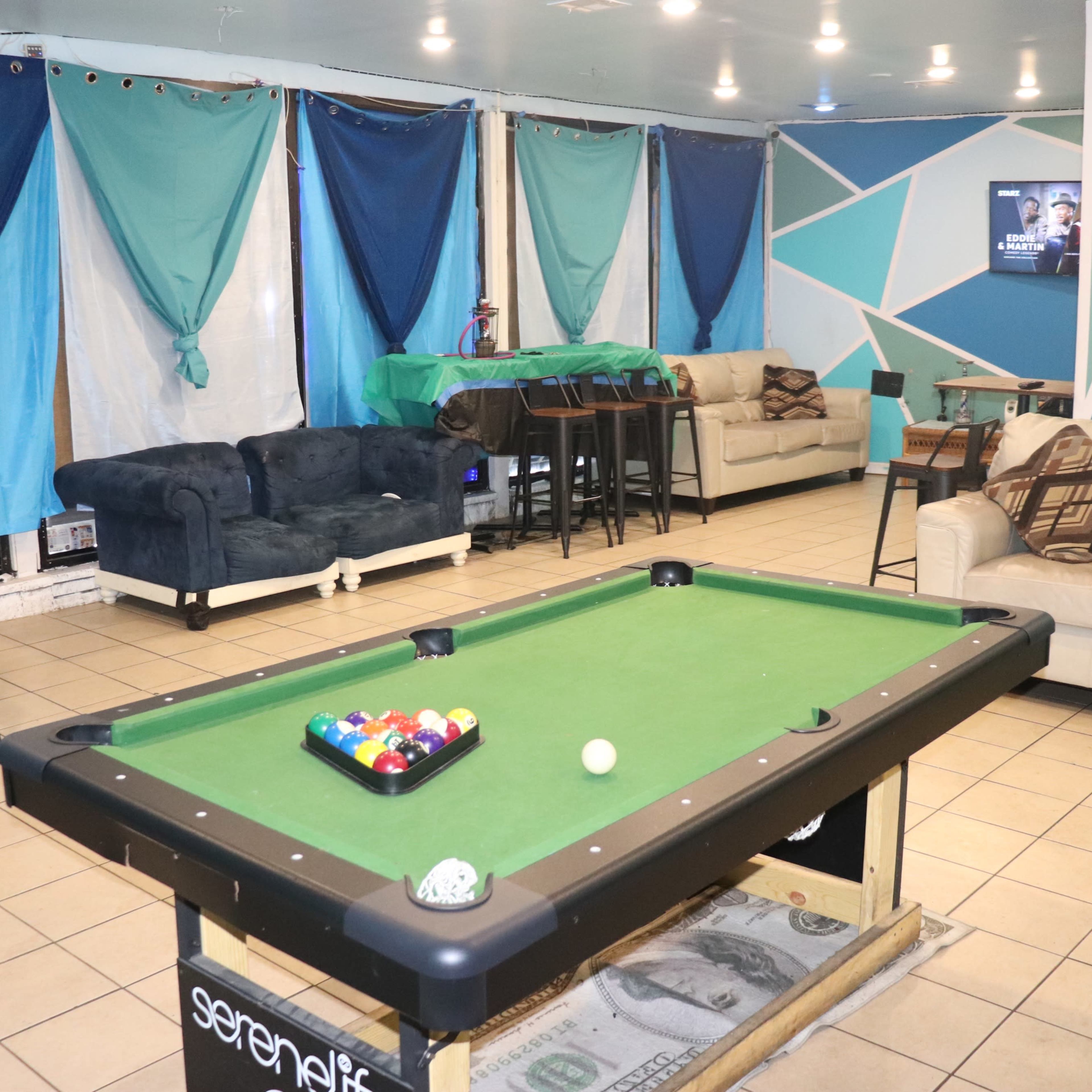 A recreation room featuring a green pool table with balls, two couches, and a table in front, all set against a backdrop of blue and teal drapes.