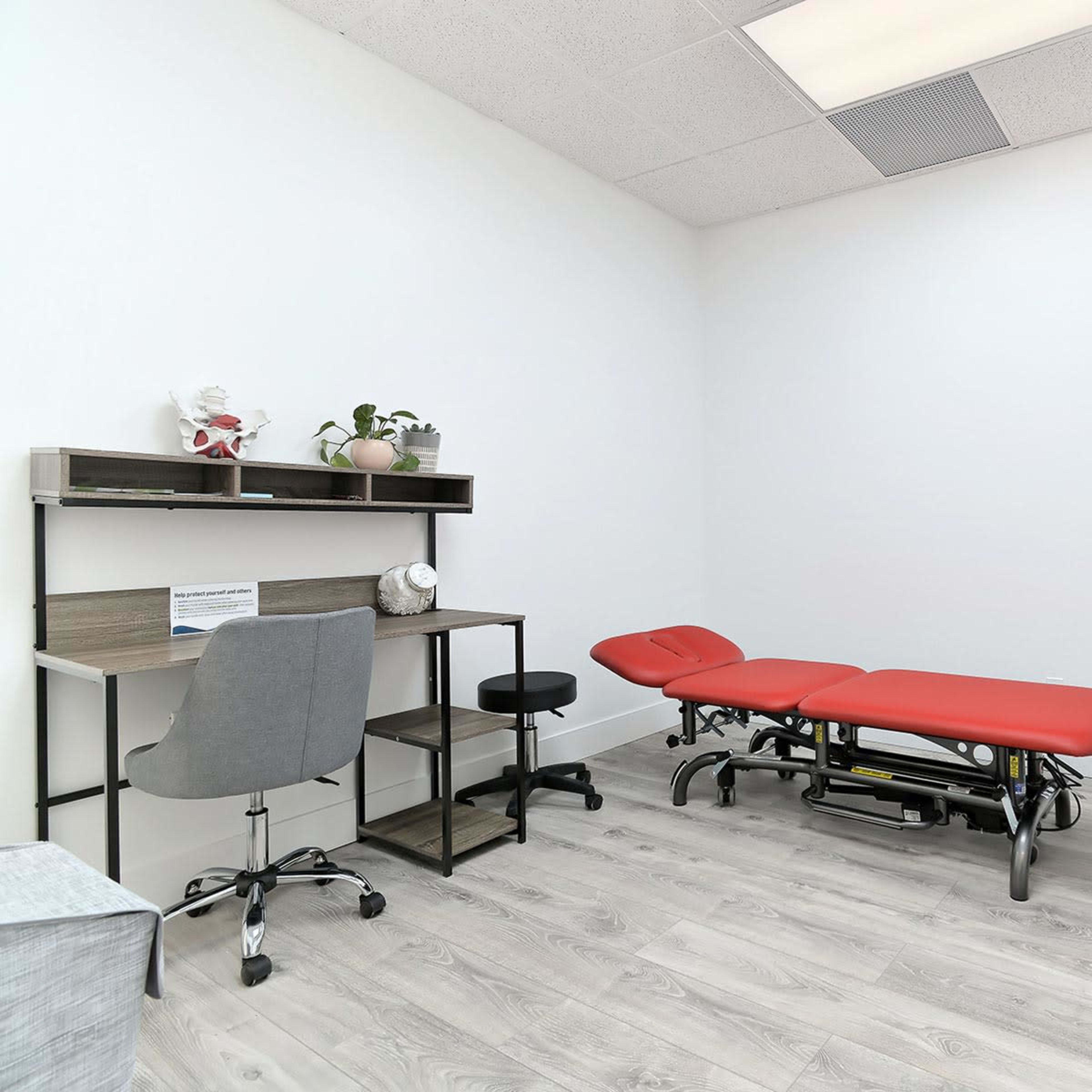 A minimalist clinical room features a red examination table, a desk with a chair, and a cabinet against a white wall.