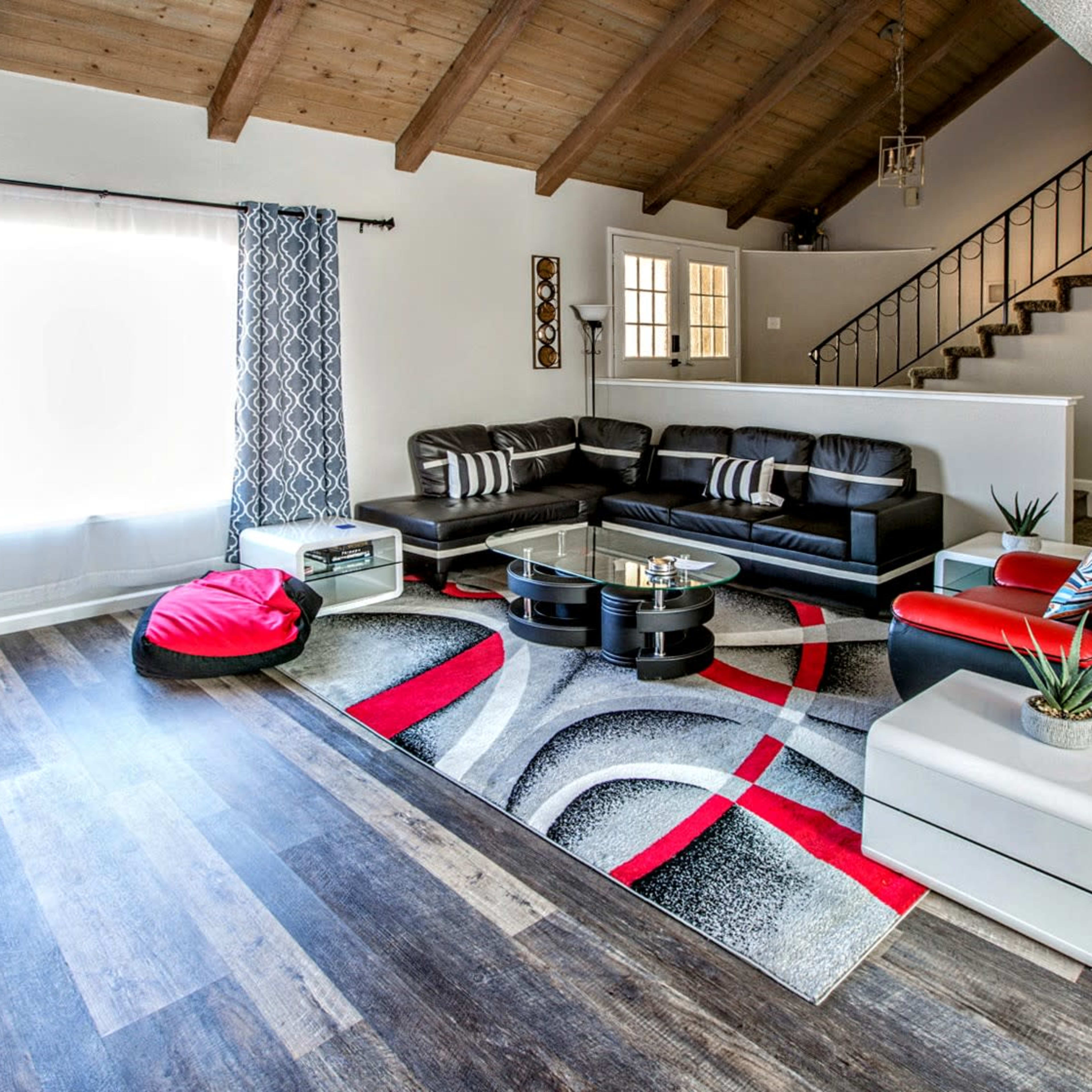 The living room features a modern black leather sectional sofa, a red accent chair, a large area rug with geometric patterns, and a high wood-beamed ceiling.