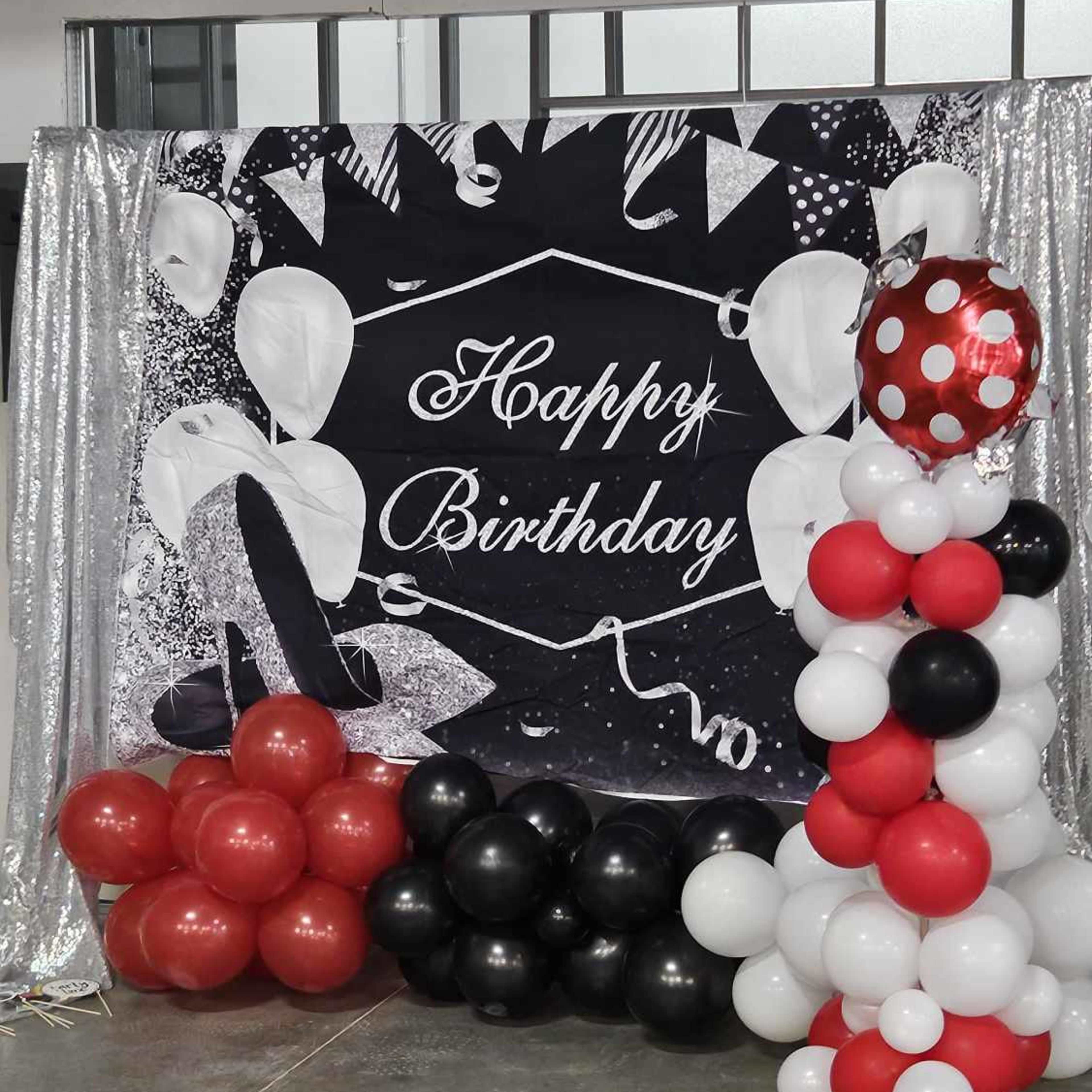 A birthday decoration featuring a backdrop with "Happy Birthday" in white text and a display of balloons in red, black, and white arranged in two clusters.