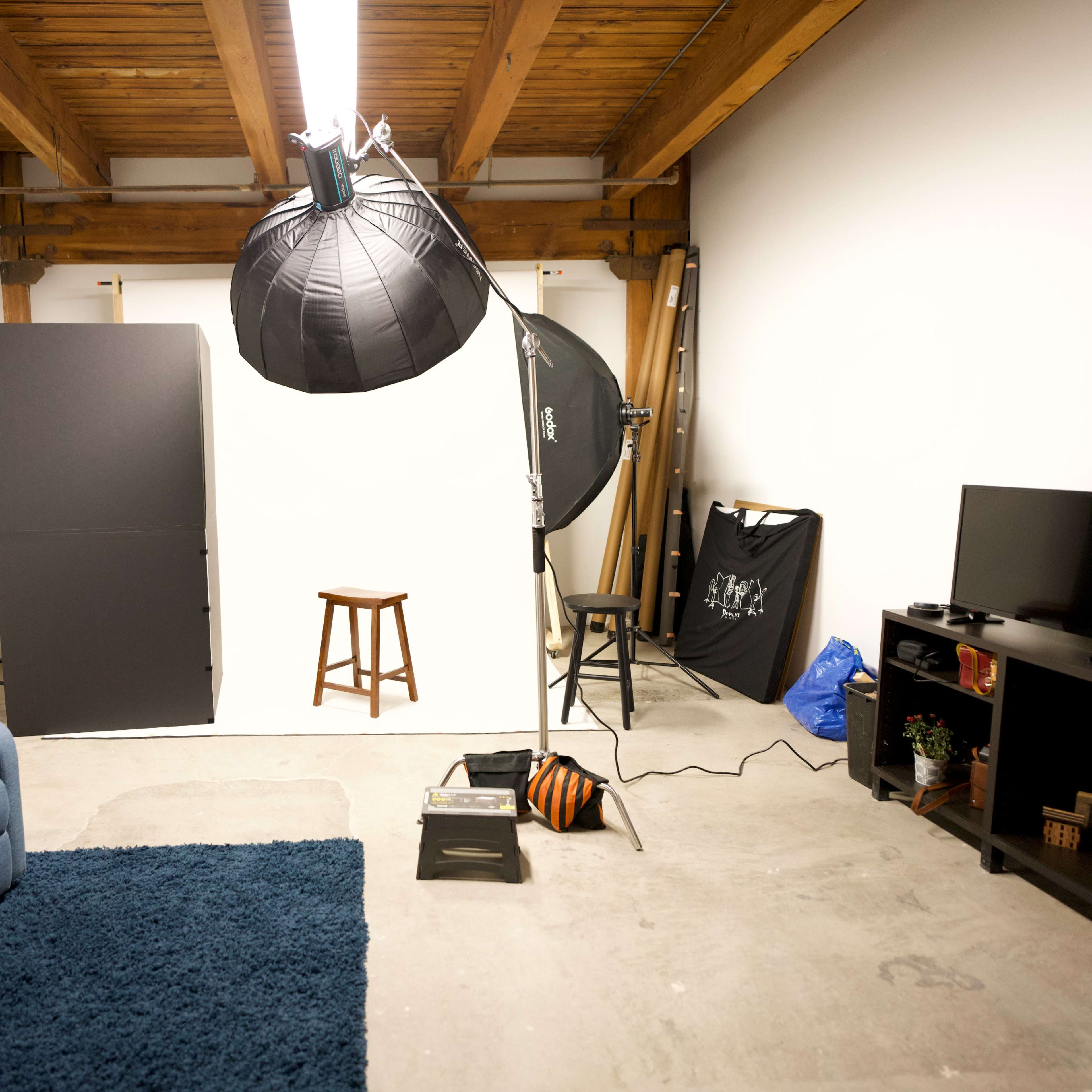A photography studio features a blue armchair, a brown stool, lighting equipment, a TV, and a backdrop in a minimally furnished space.