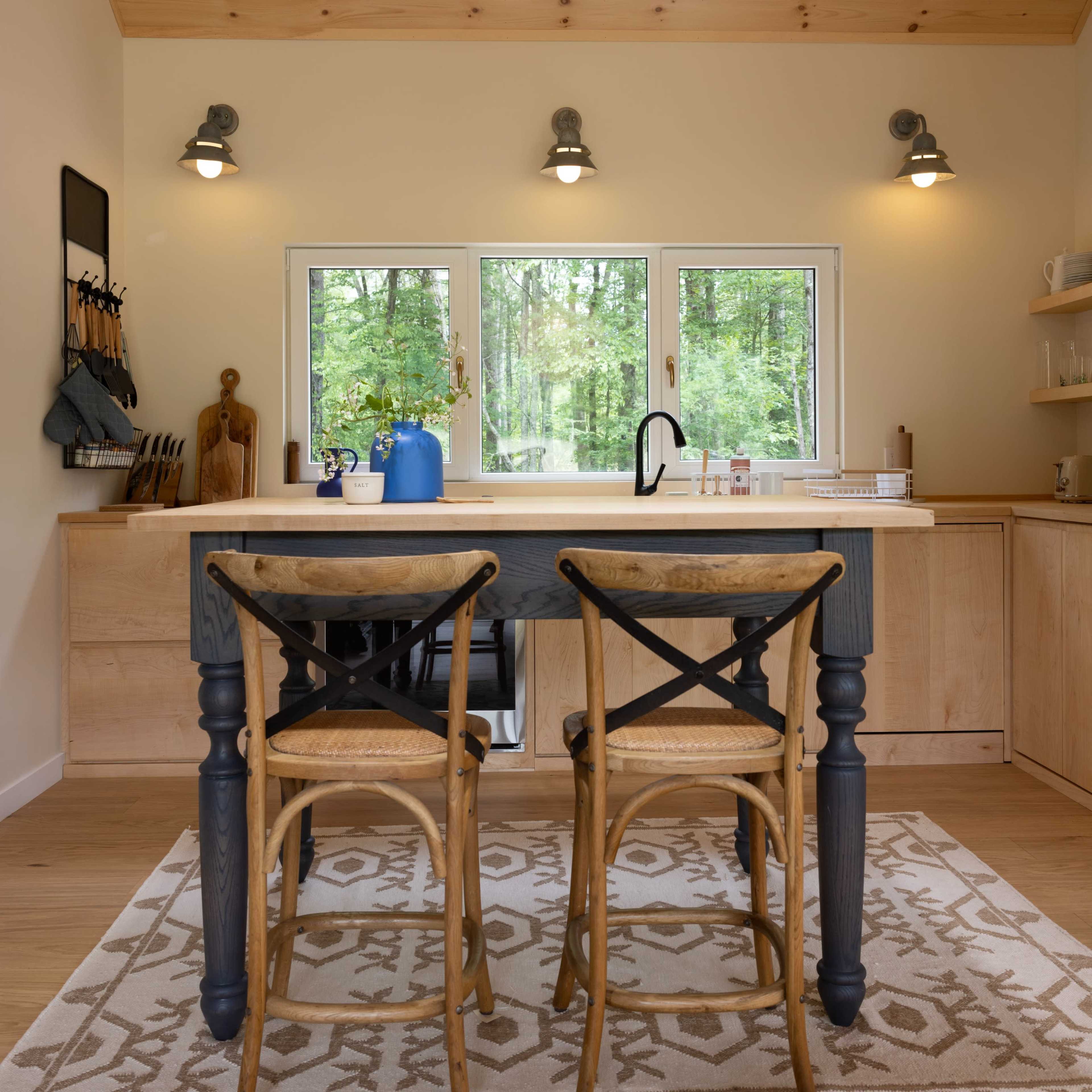 A modern kitchen features a wooden countertop with two beige bar stools, pendant lighting, and large windows overlooking a forested area.