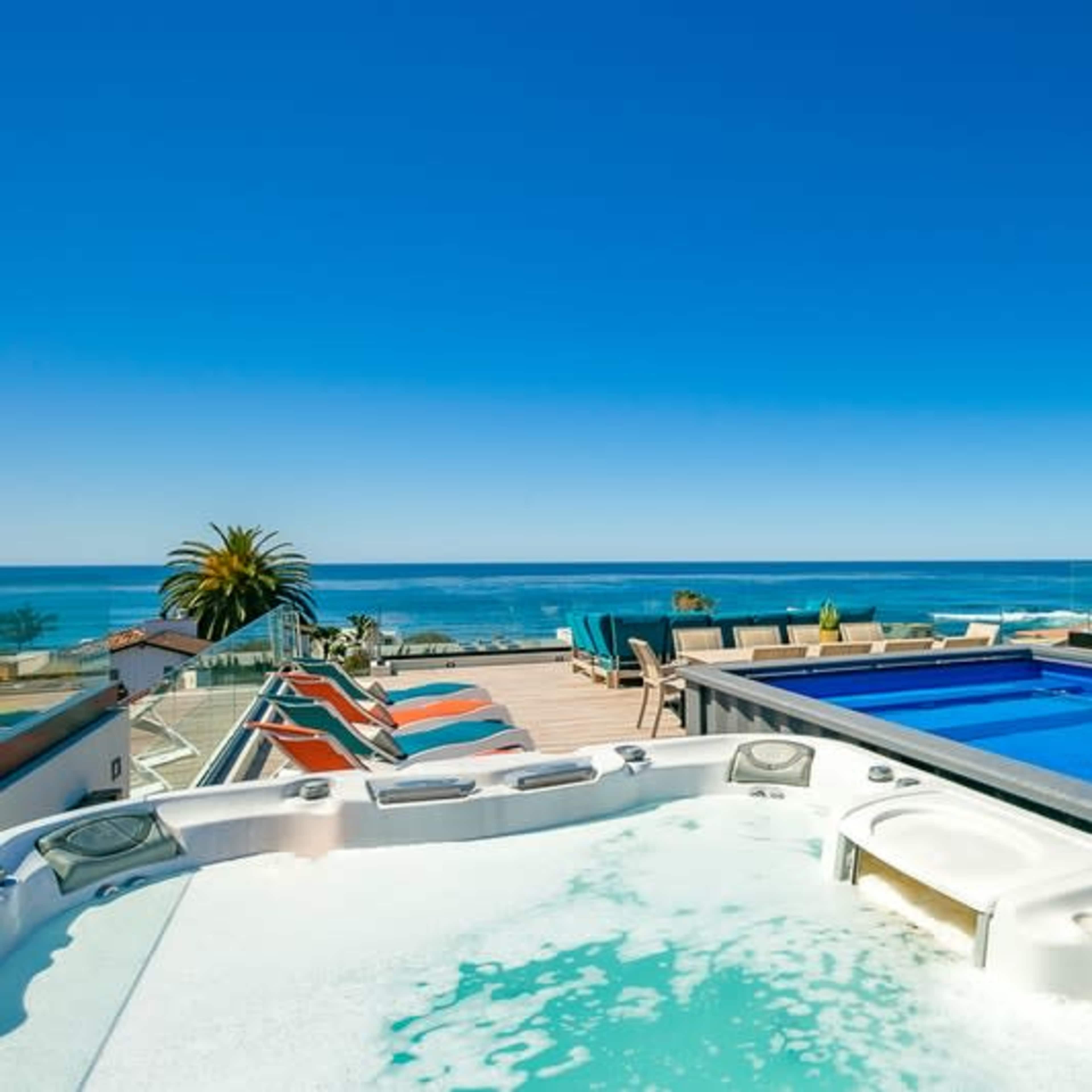 A hot tub sits in the foreground next to an infinity pool overlooking the ocean under a clear blue sky.