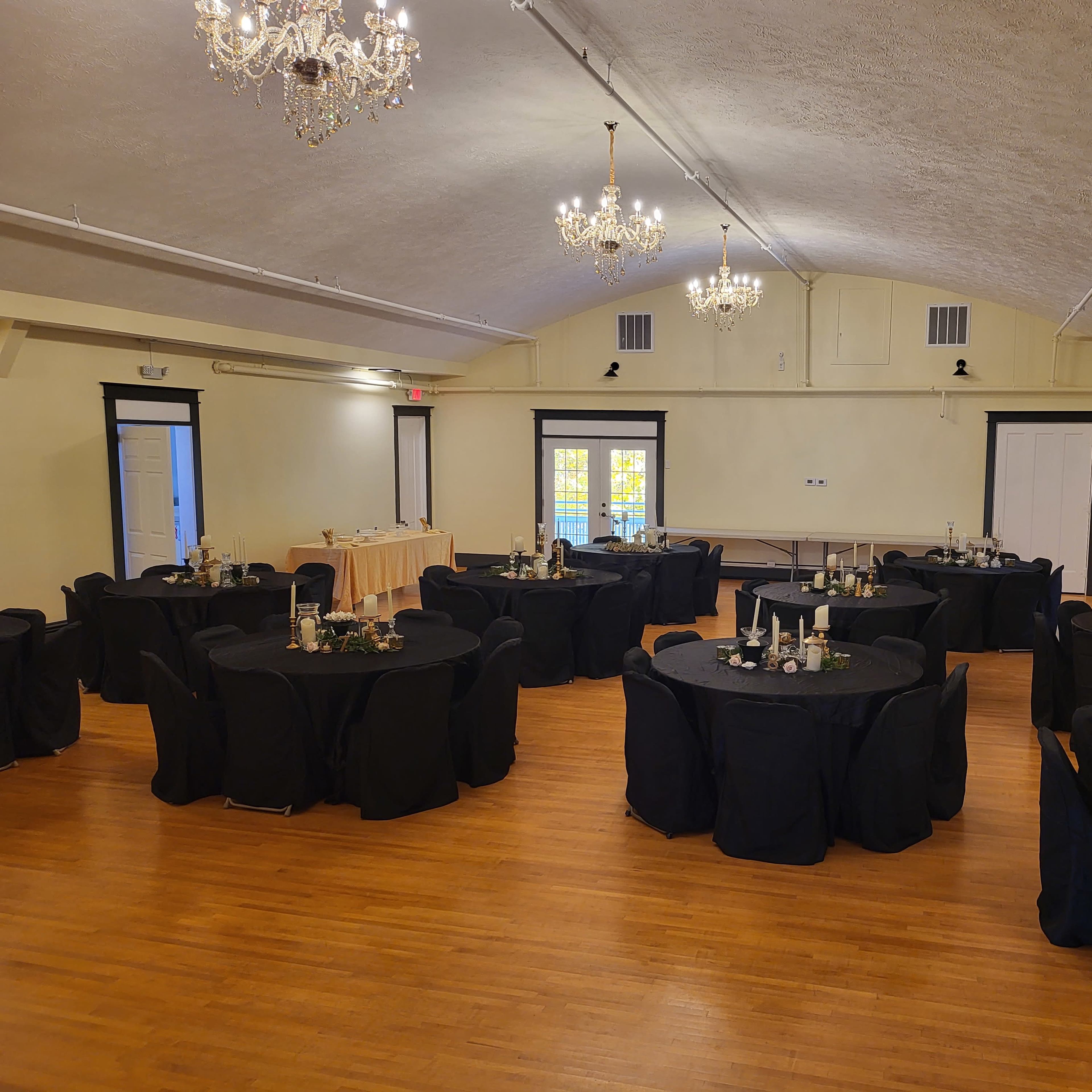 A large event space features several round tables covered in black tablecloths, each adorned with centerpieces, under a ceiling with chandeliers.