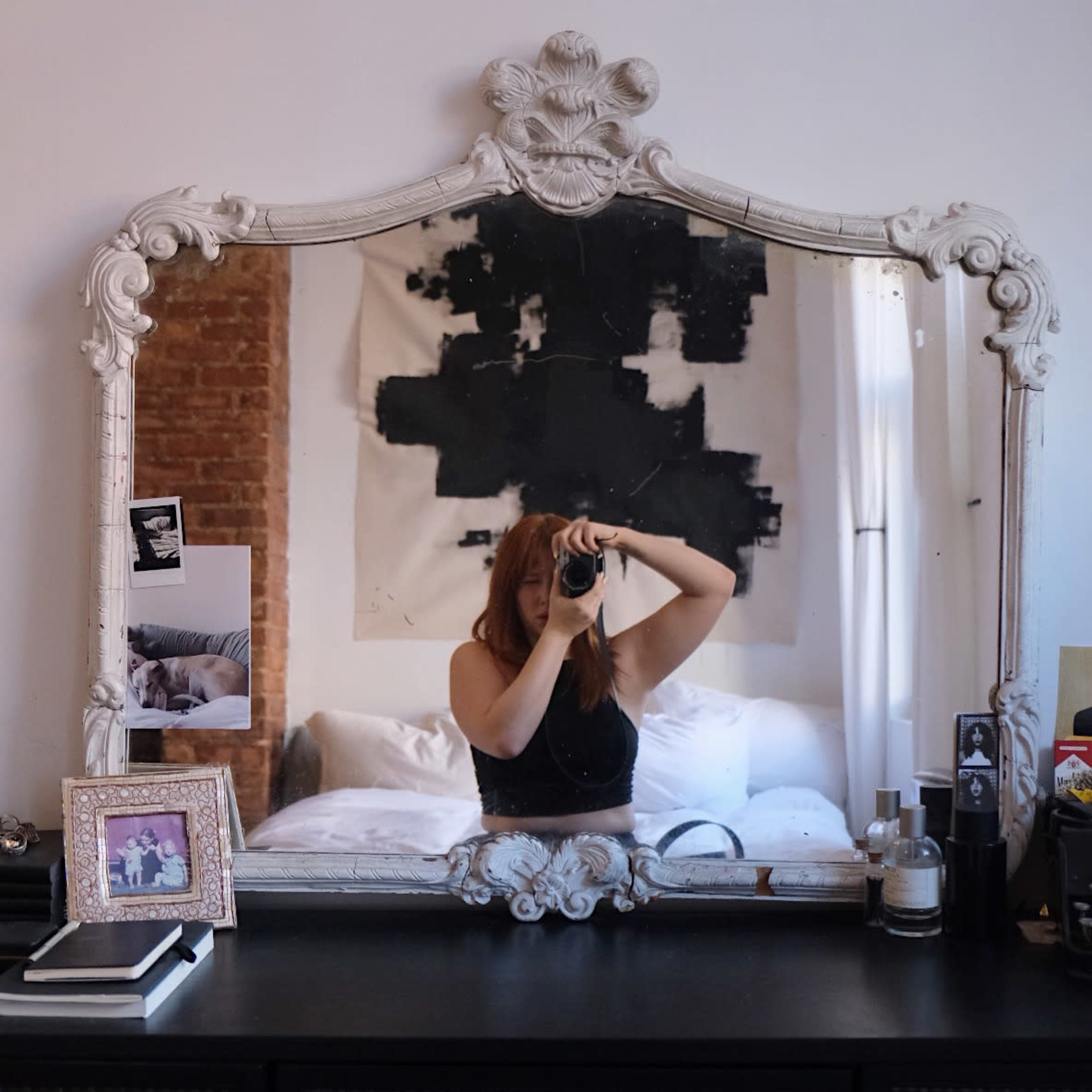 A person takes a picture of themselves in a mirrored frame above a dark dresser in a room with a patterned wall.