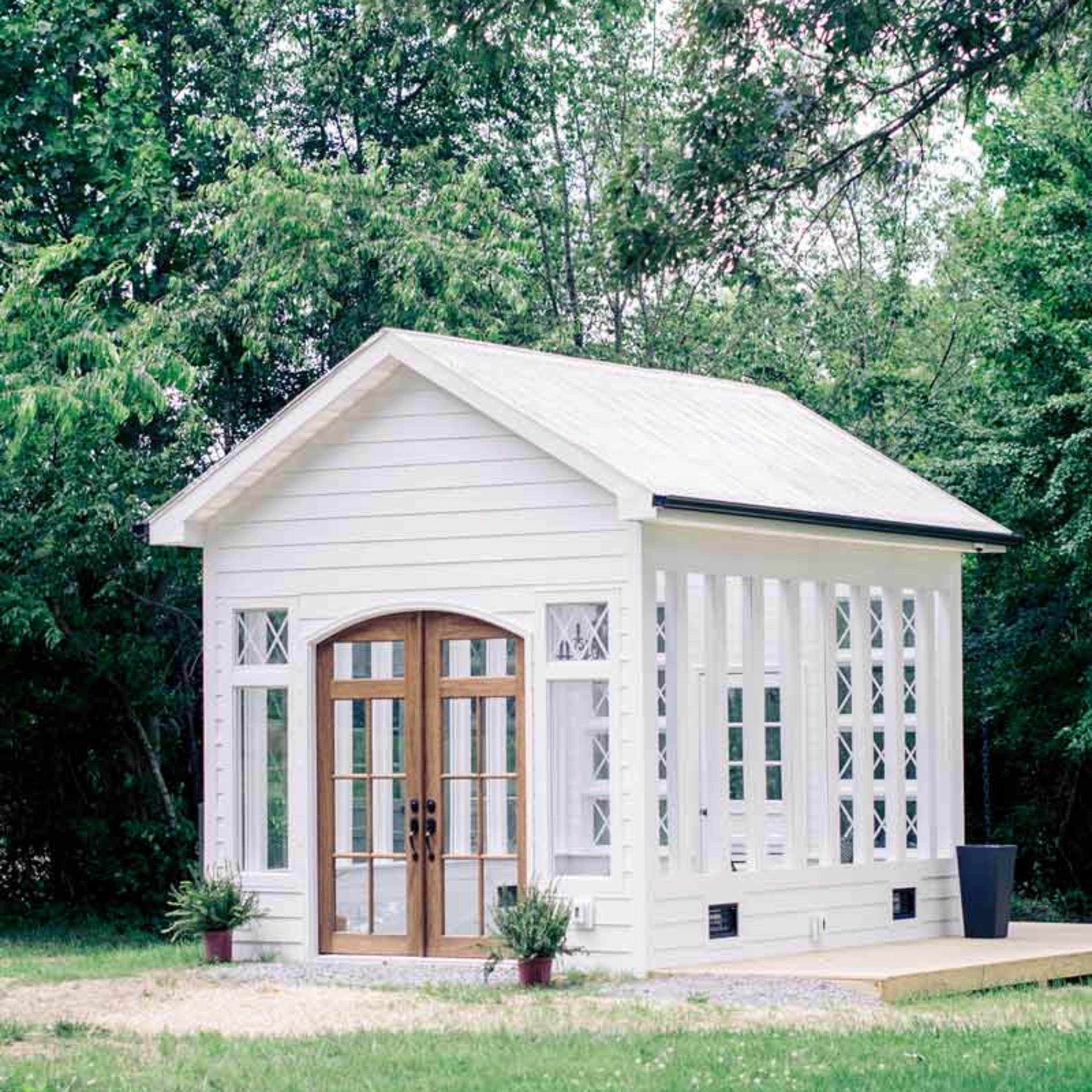 A small white structure with large glass doors and windows is surrounded by greenery and sits on a grass lawn.