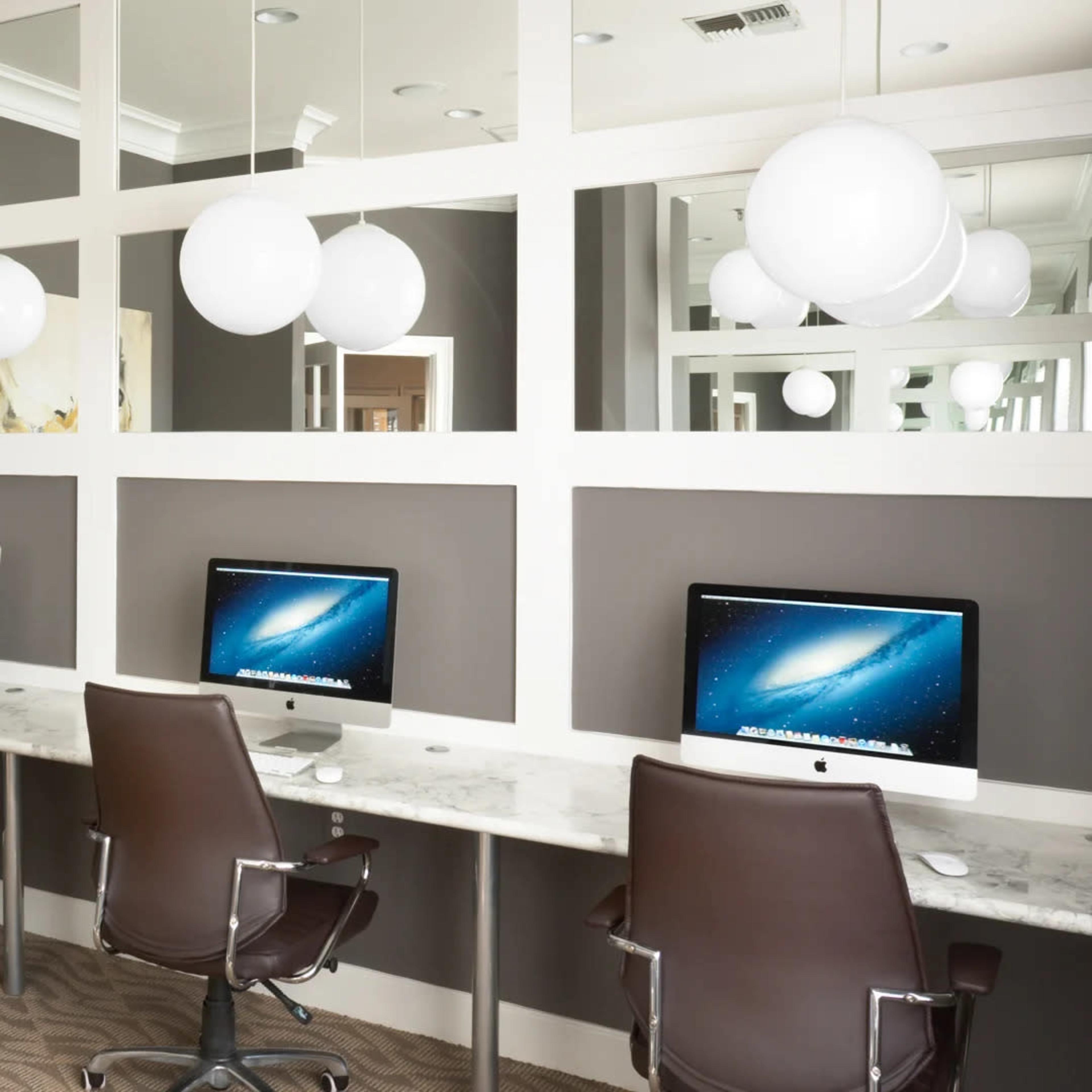 A modern office space with three computer workstations, each equipped with an iMac, facing a large, abstract painting and illuminated by pendant lights.