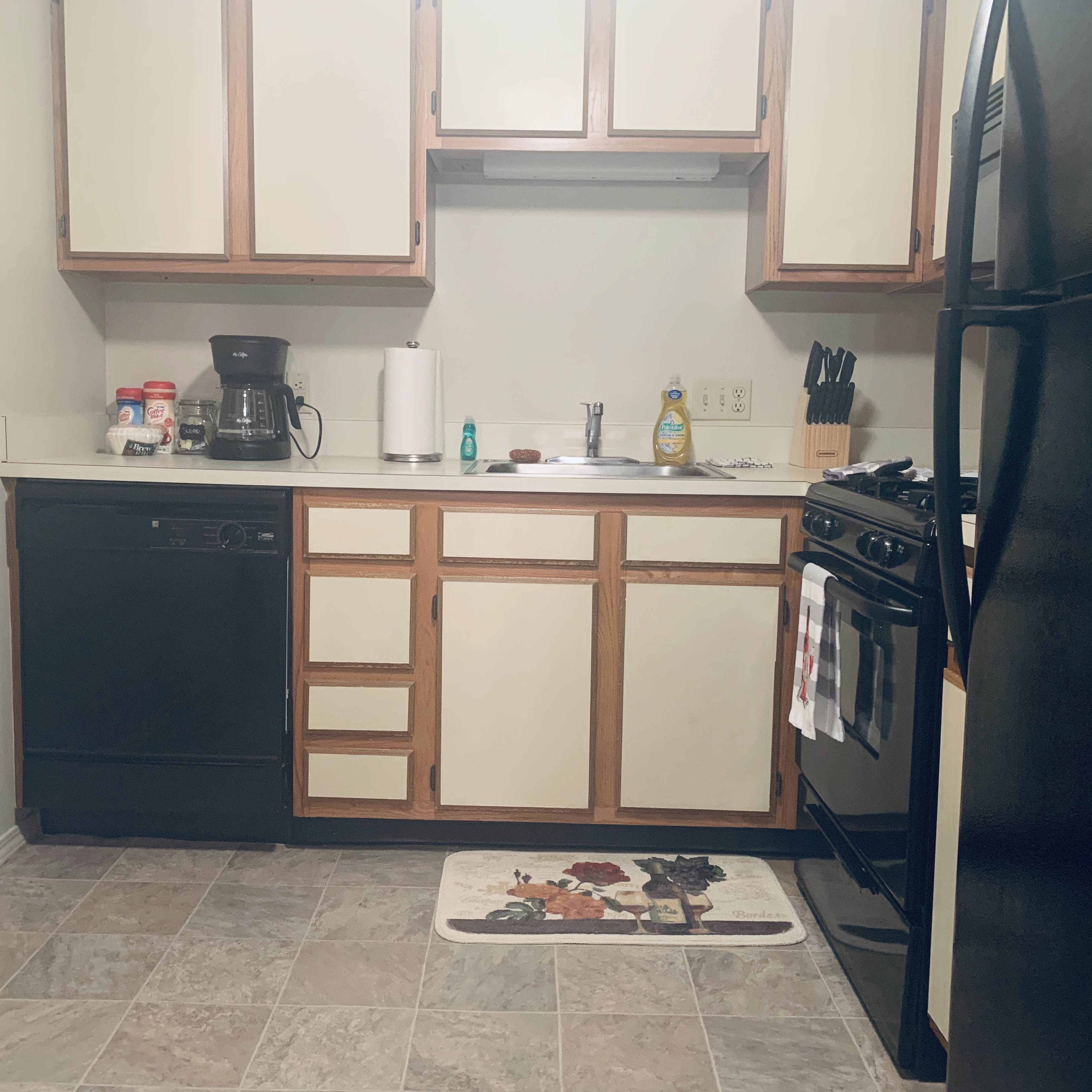 A kitchen with light-colored cabinets, black appliances, and a tiled floor is shown, featuring a coffee maker, dish soap, and a dish towel.