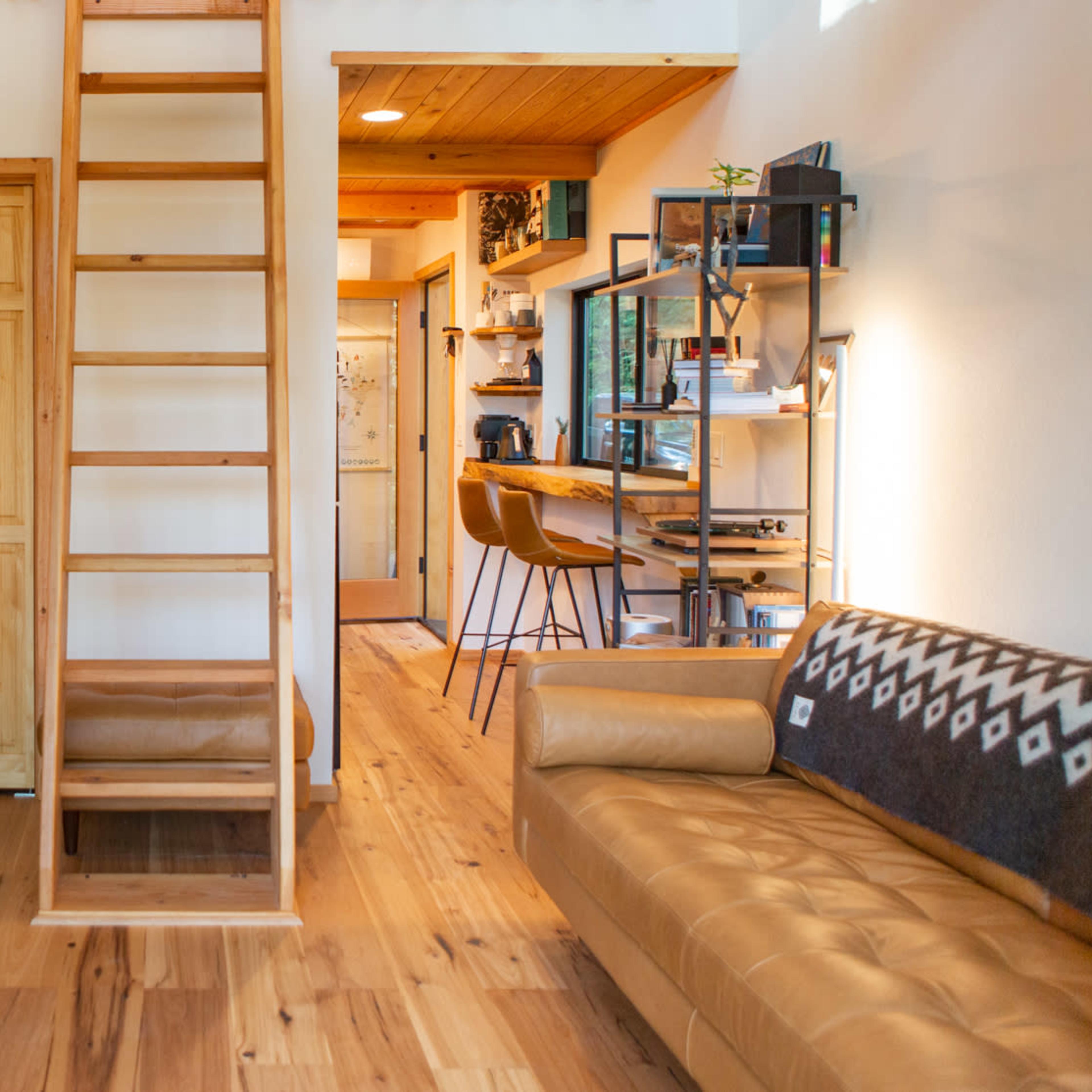 A small living space featuring a wooden ladder leading to an upper area, a desk with a computer, a sofa, and a kitchen area with stools.