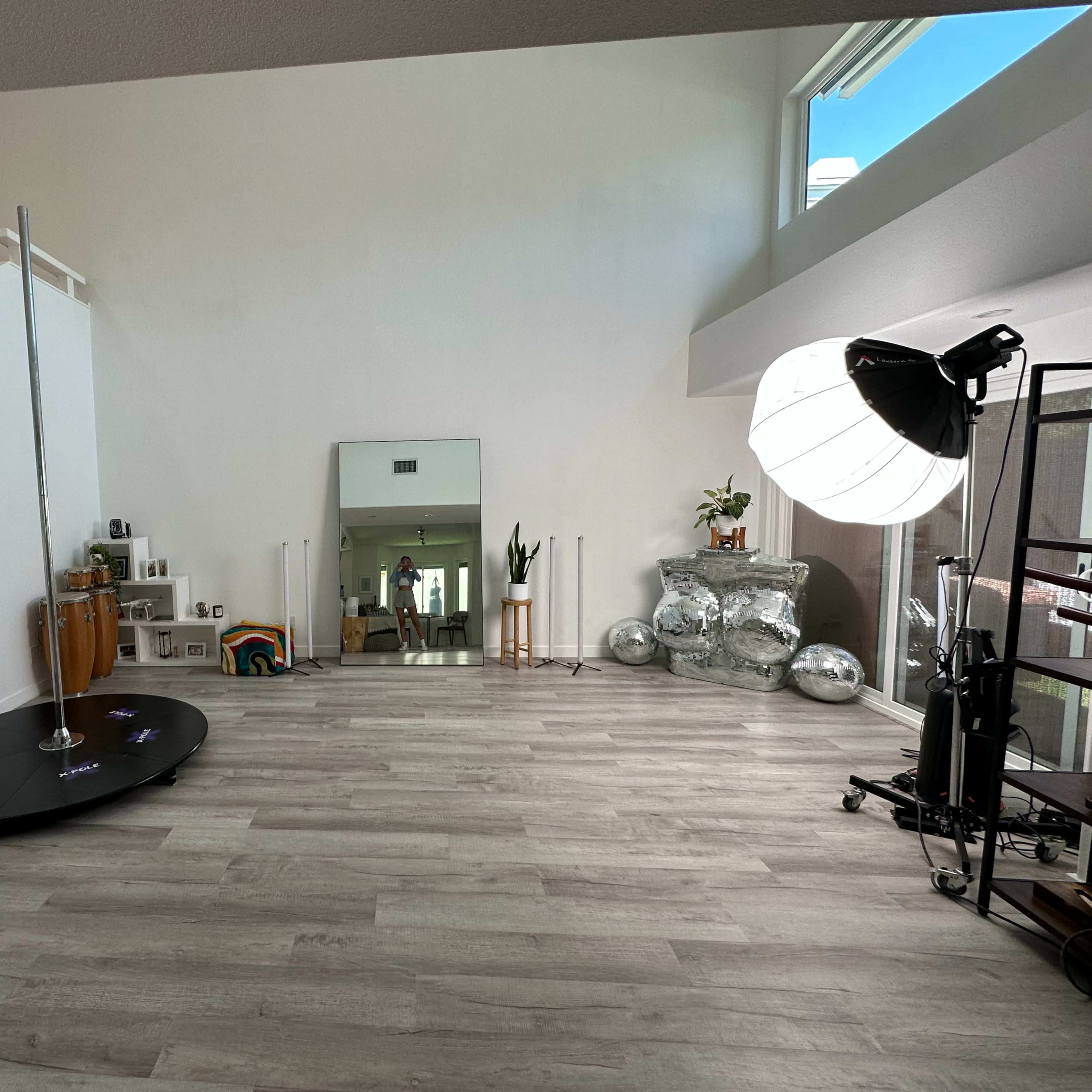 A spacious, modern room with light gray flooring, featuring a pole, a large mirror, a reflective object, and a photography light setup.