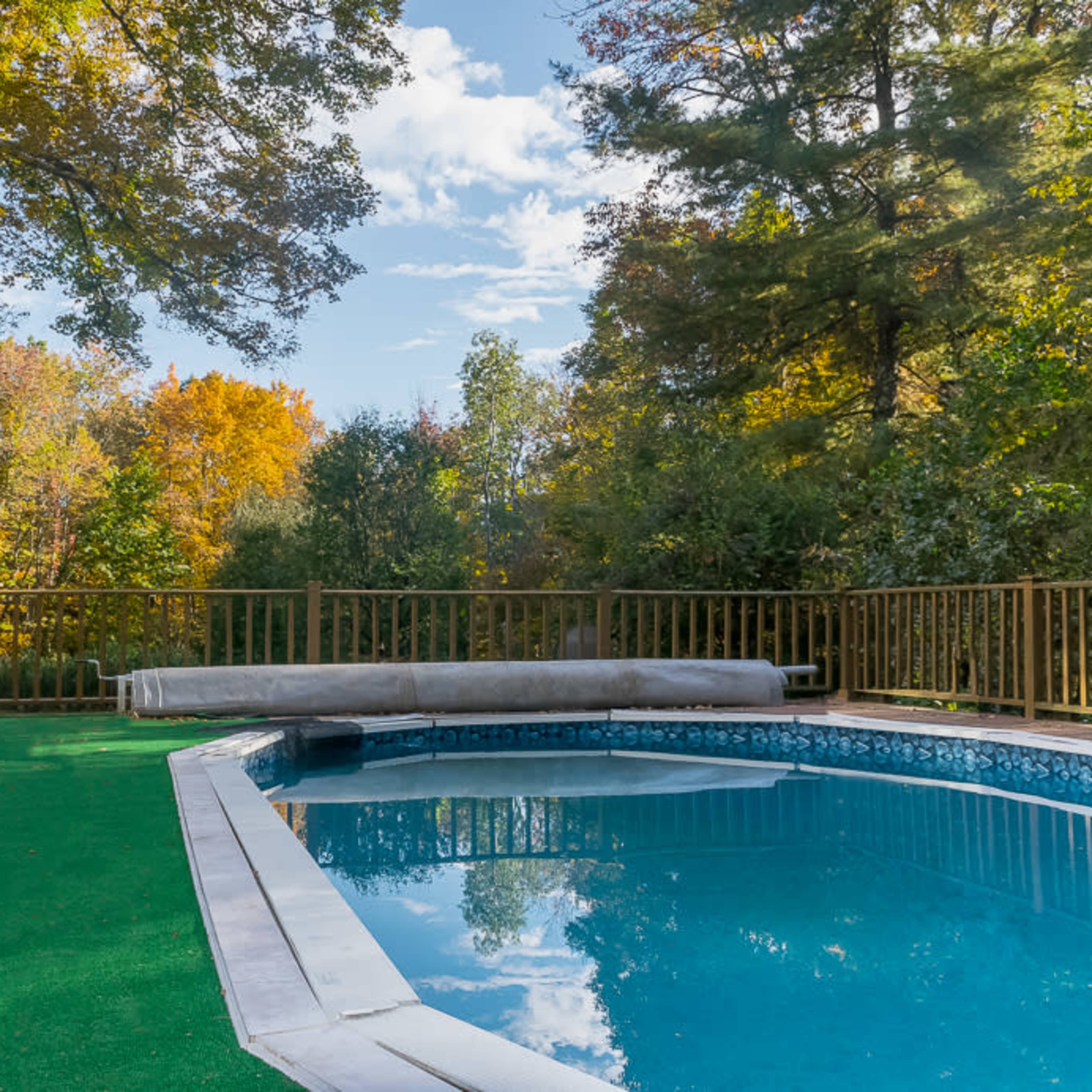 A swimming pool surrounded by a wooden deck and green lawn, with trees displaying fall foliage in the background.